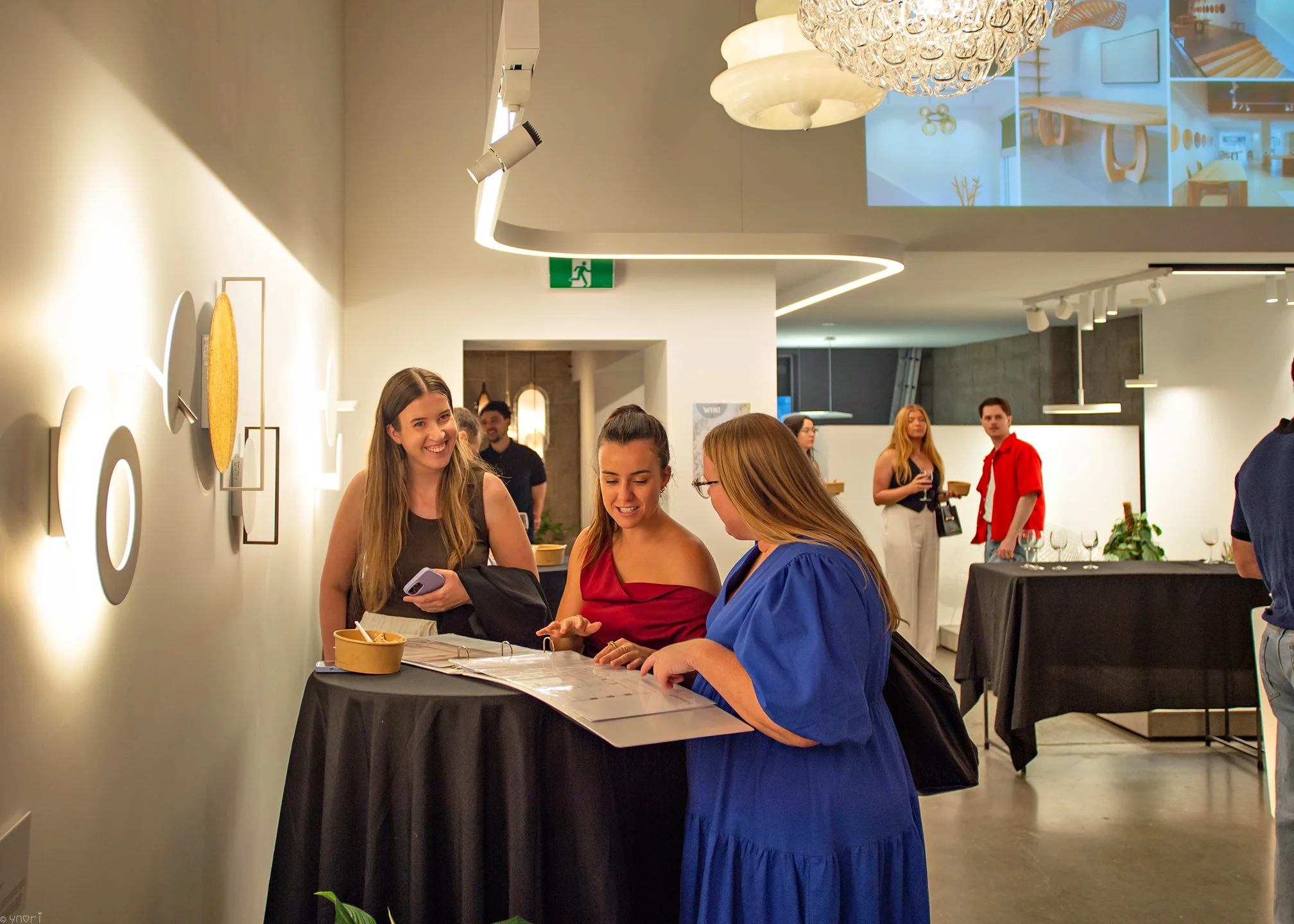 Group of women at a reception desk in a modern indoor event space, engaging with a woman in a blue dress, with others in the background near a table with wine glasses and a projection of interior designs on the ceiling.