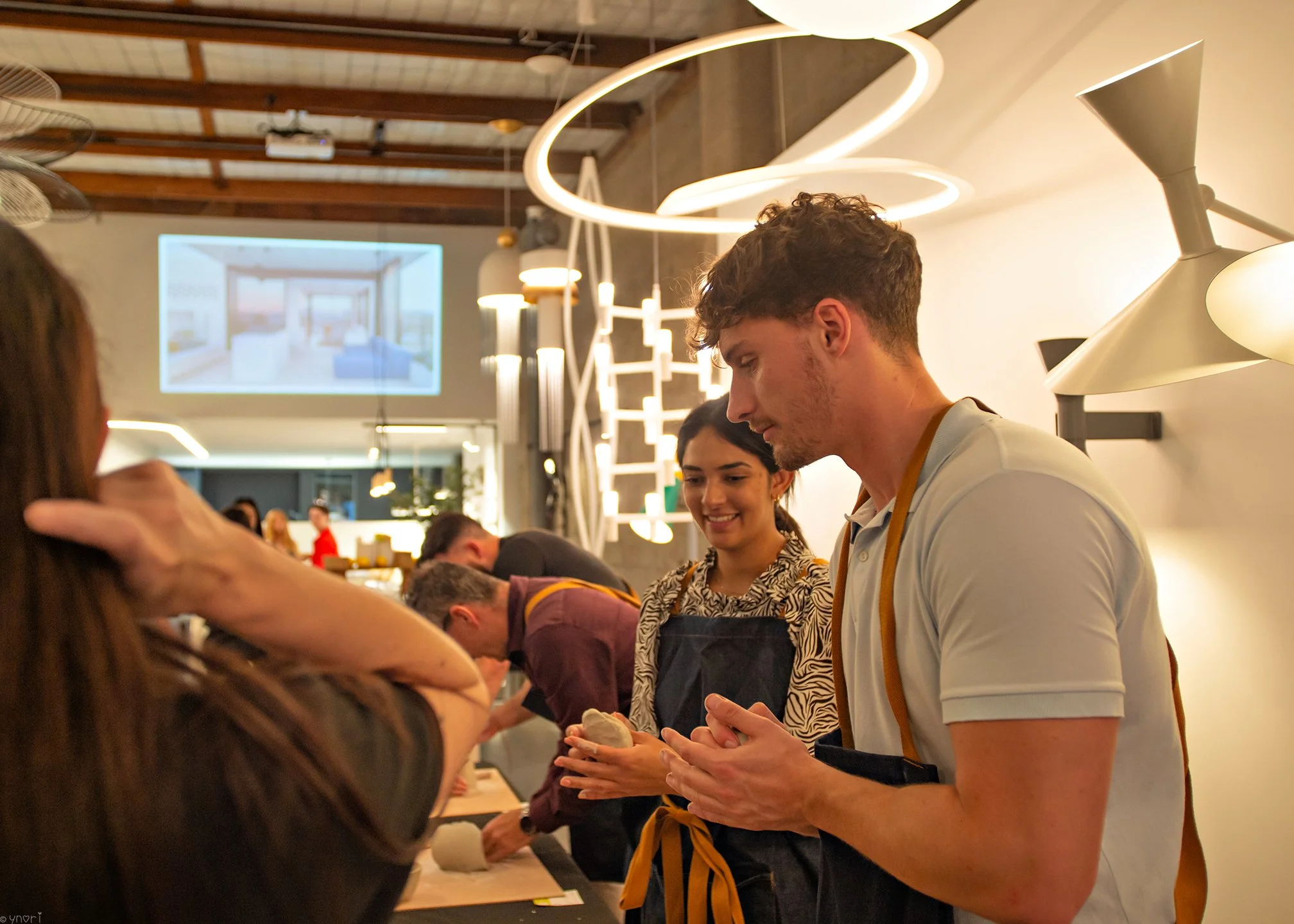 A group of people, including a young man and woman, participating in a cooking class or demonstration in a modern kitchen with bright lighting and contemporary decor.
