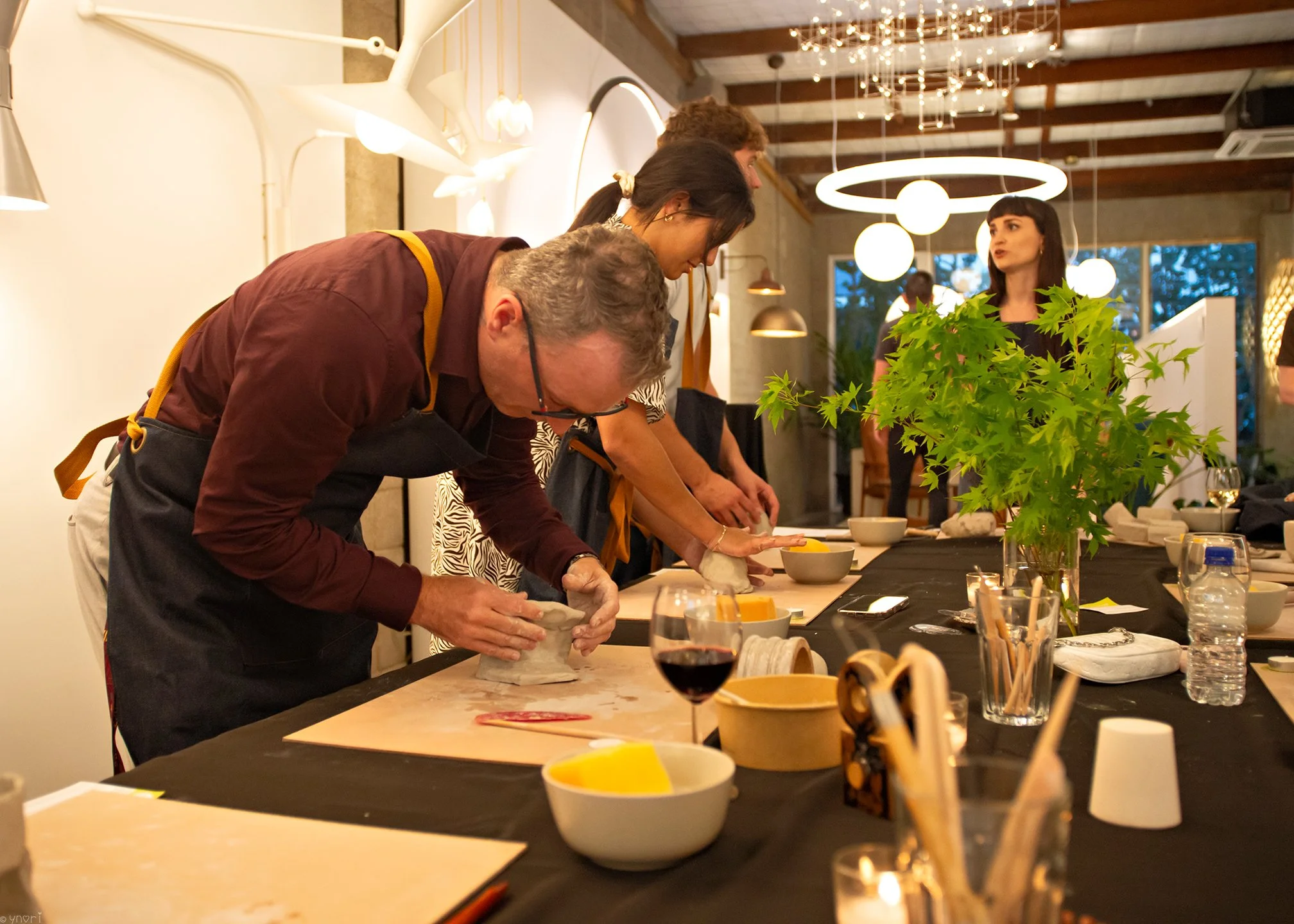 People participating in a pottery class or workshop, working on clay projects at a table in a well-lit room with decorative hanging lights and a plant in the center.