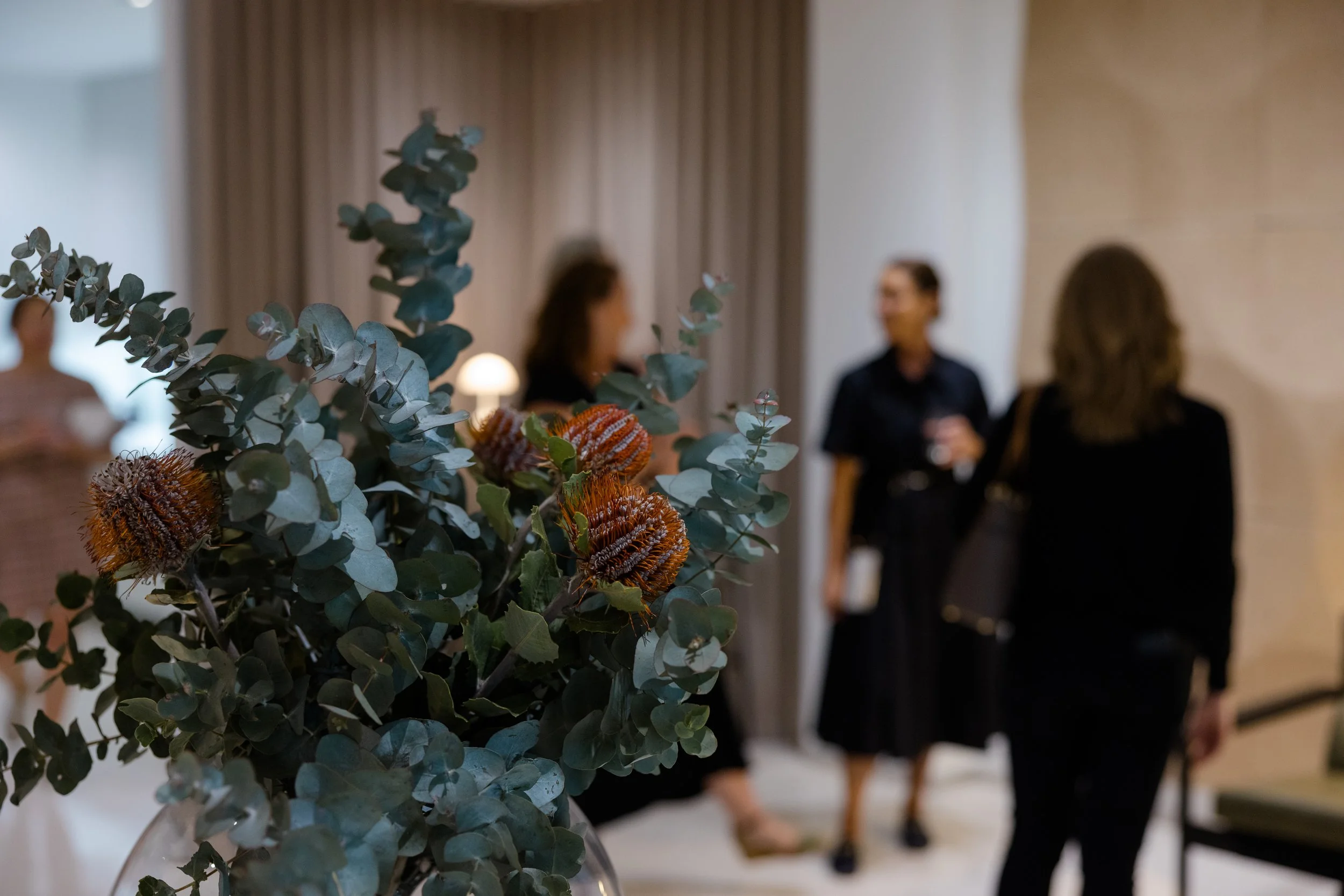 Close-up of a floral arrangement with eucalyptus leaves and orange flowers, with three women in the background talking indoors.