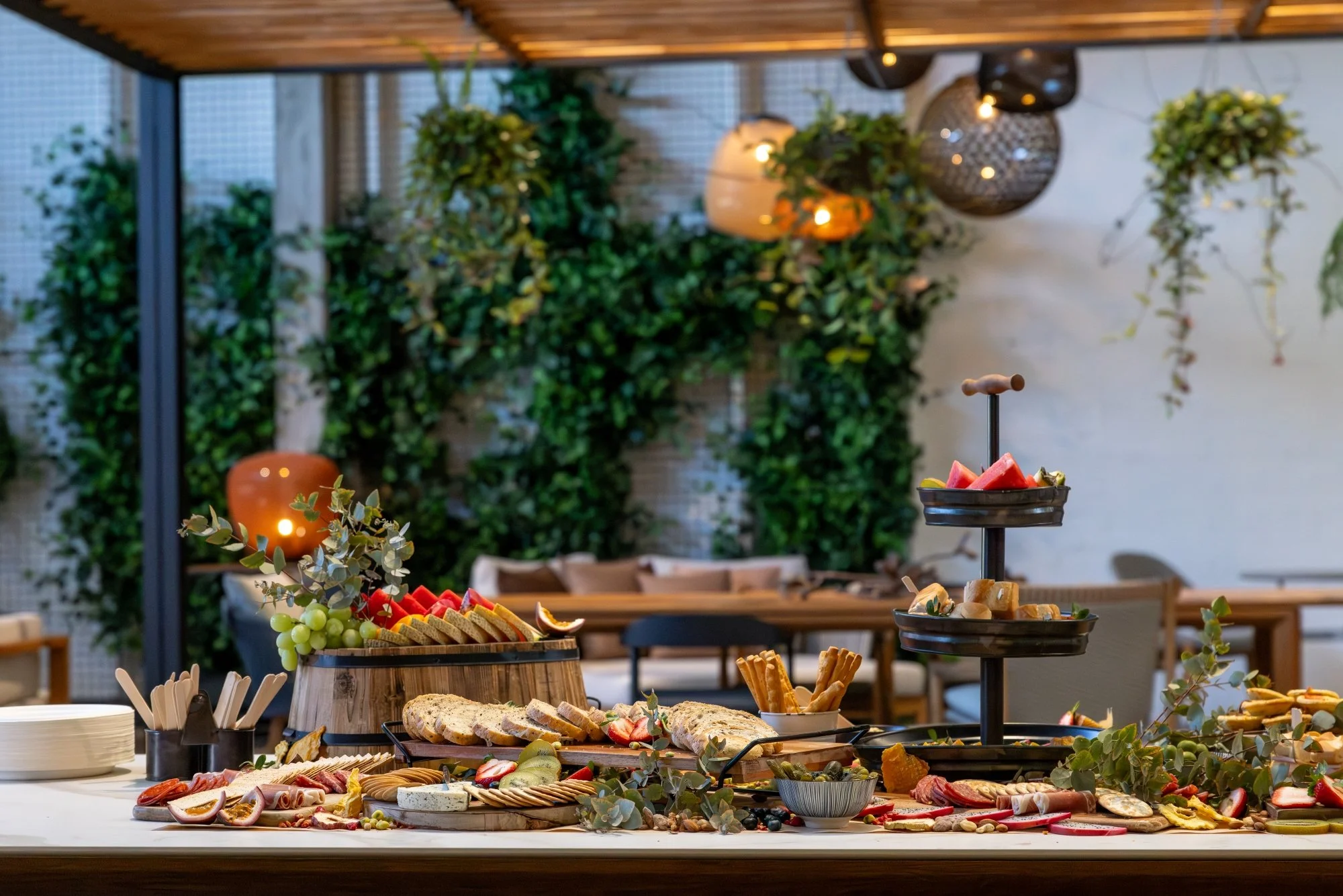 A buffet table with assorted snacks, sliced bread, fruits, crackers, and vegetables in a decorated indoor setting with green plants and hanging lights.
