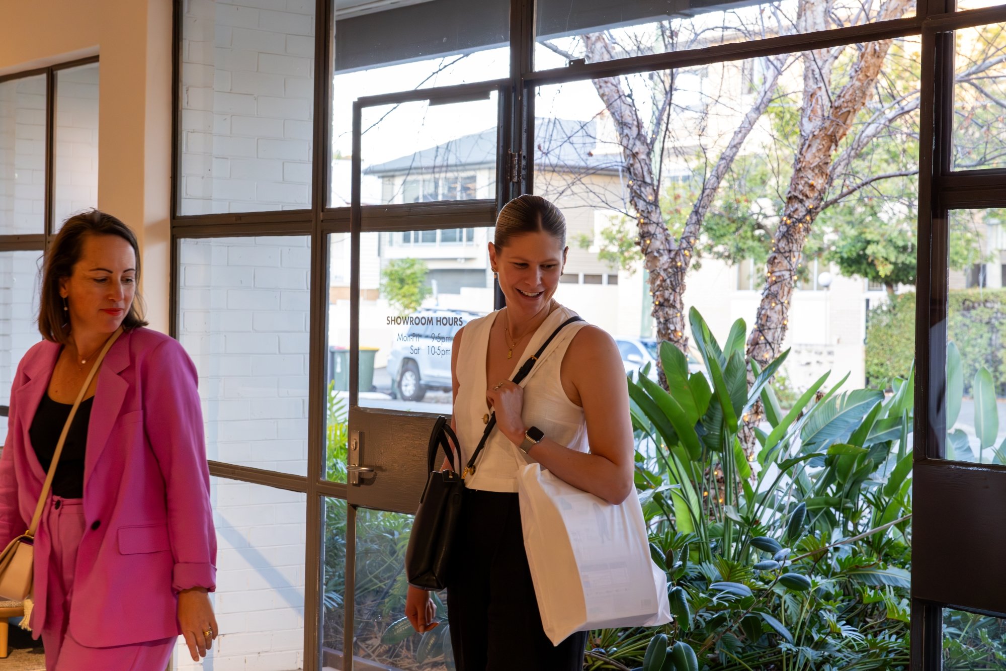 Two women entering a building with large glass windows and greenery outside. One woman is in a pink suit, the other in a white sleeveless top holding a tote bag.