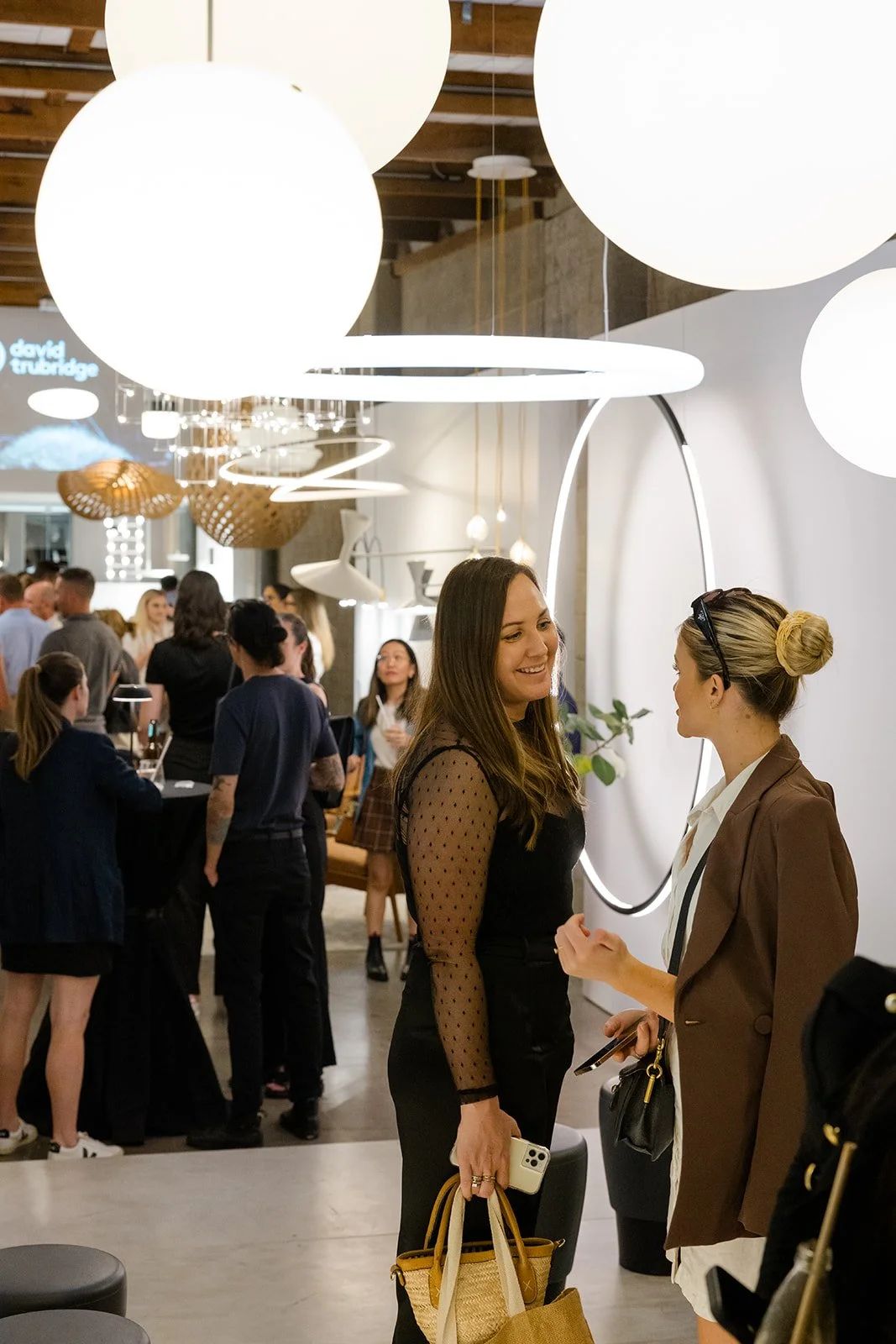 Two women are conversing in a modern, well-lit interior space filled with light fixtures and a crowd of people in the background.