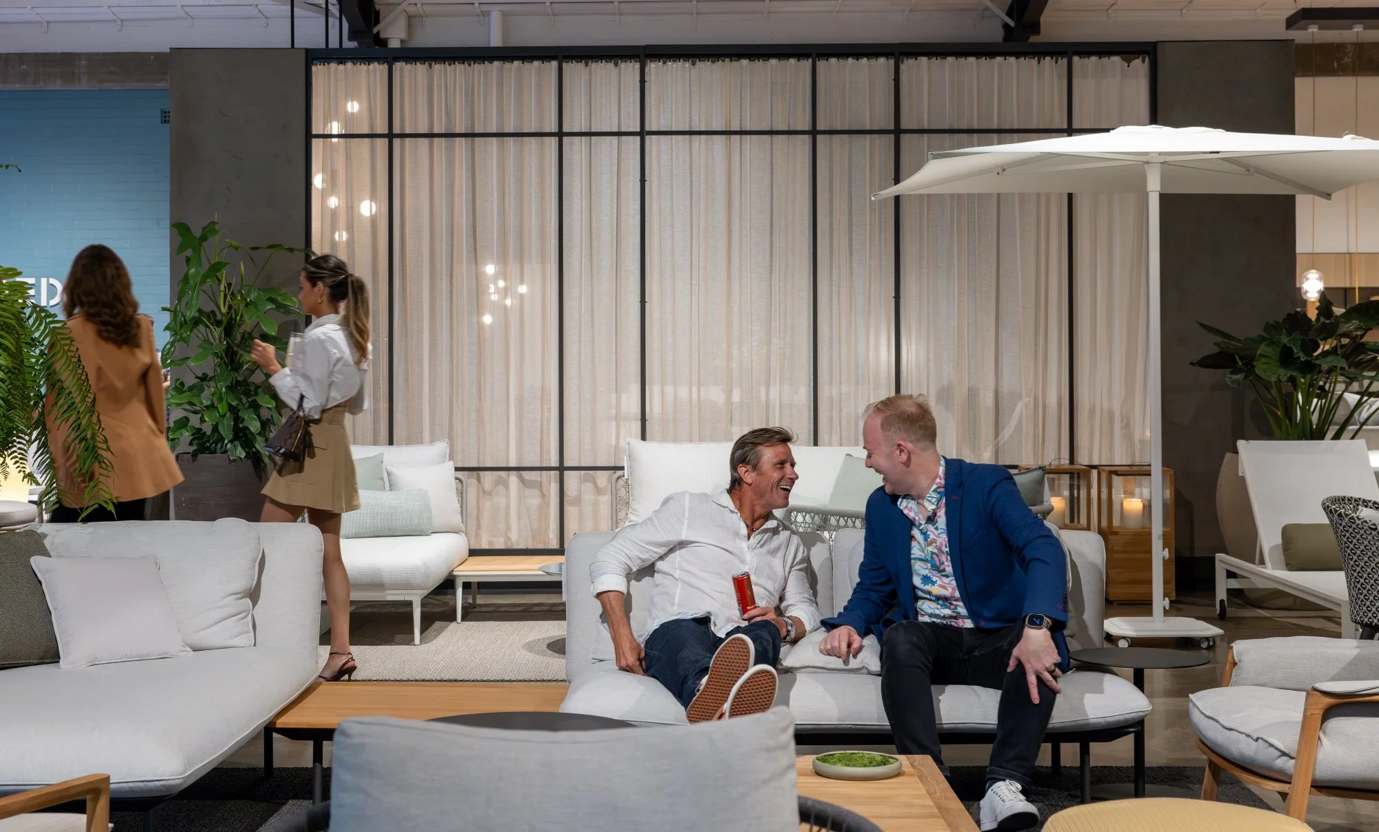 Two men sitting on a sofa in a modern lounge, smiling and talking, with two women walking by in the background. One man is holding a red object, and the other is wearing a colorful shirt and navy blazer.