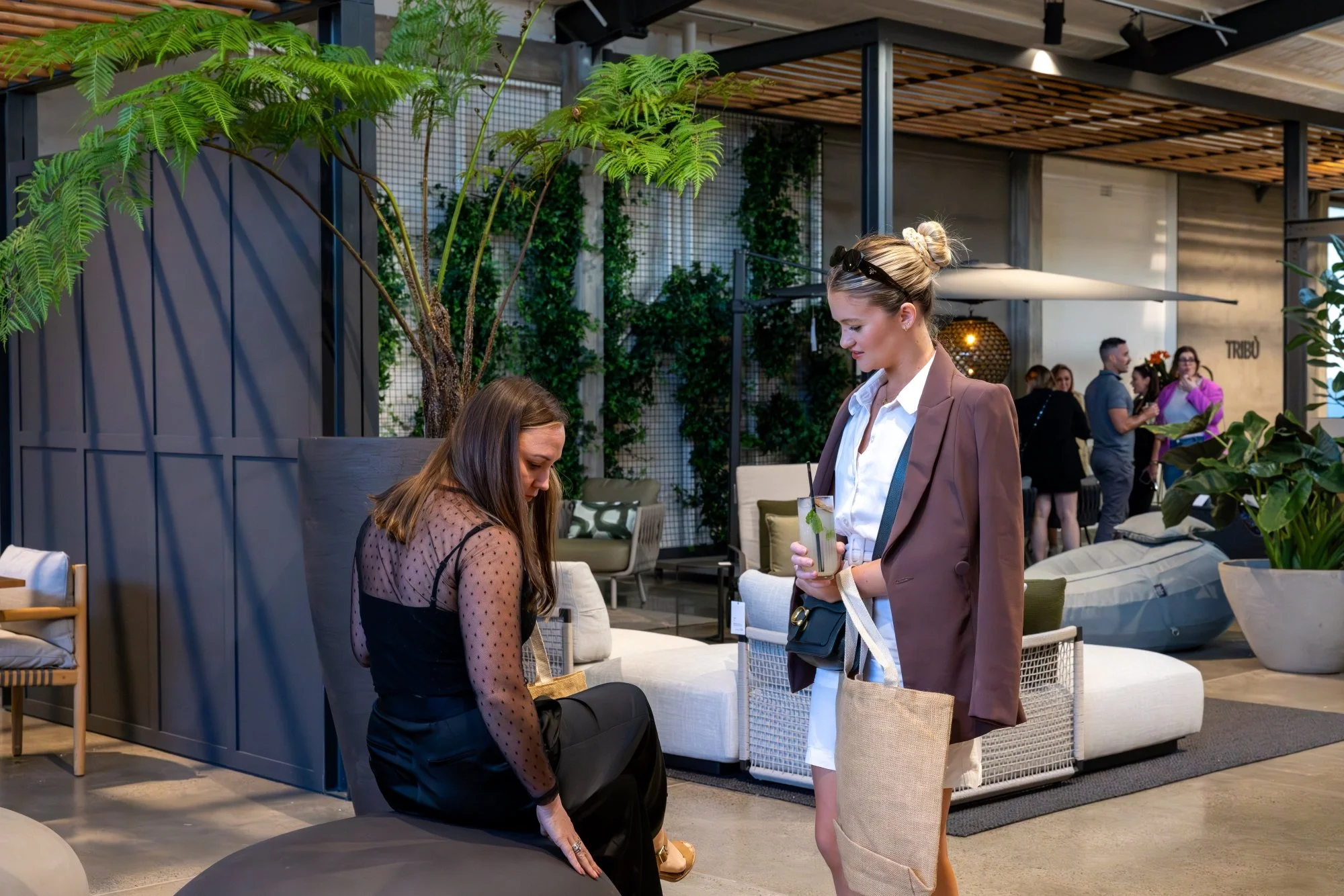 Two women in a modern indoor lounge; one is seated, adjusting her shoe, and the other is standing with a drink and a tote bag, engaging with the seated woman. There are plants, sofas, and a few people in the background.