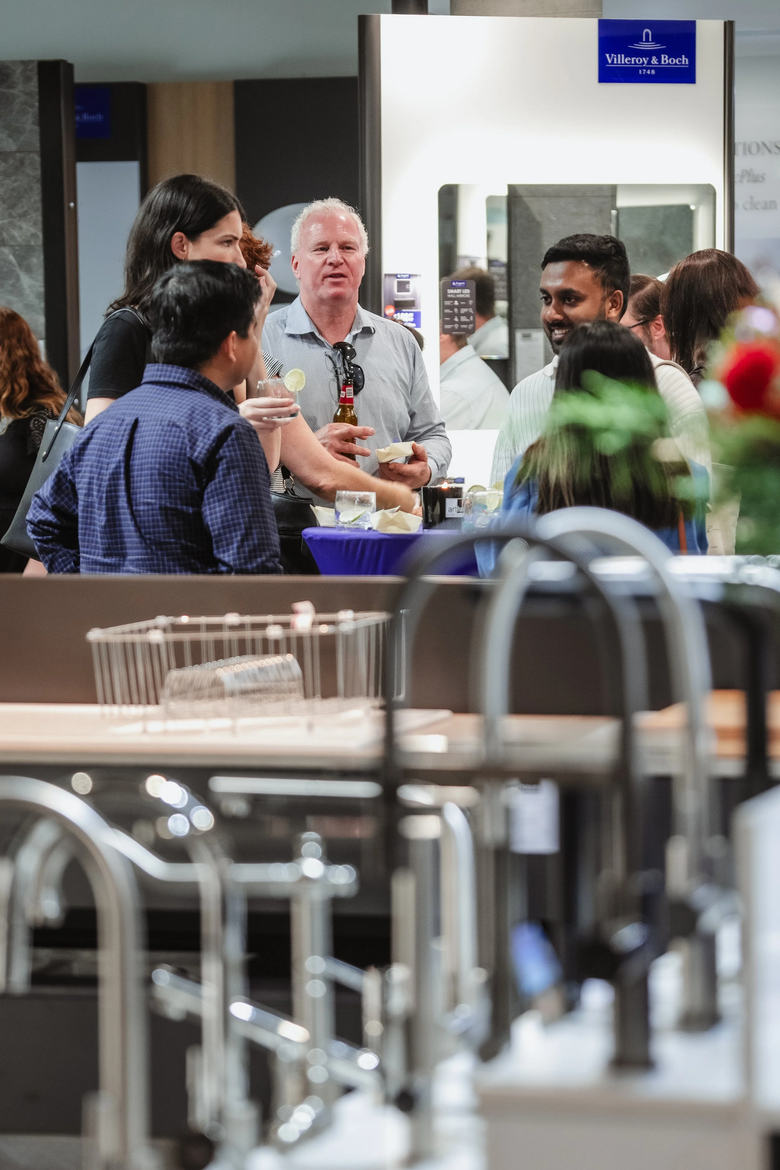 Group of people socializing at a food court or exhibition, with a heating or distribution area in the foreground and a Villeroy & Boch sign in the background.