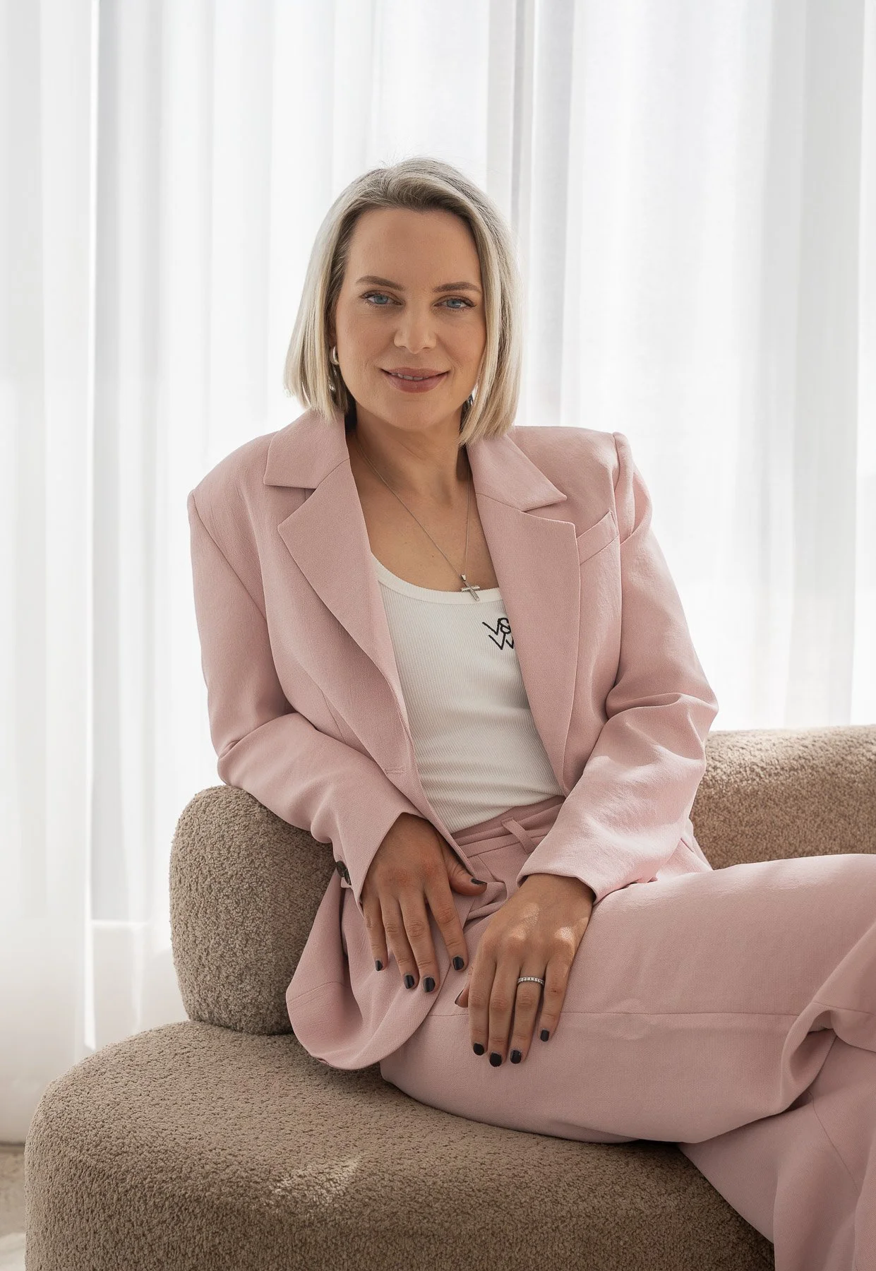 A woman with blonde hair sitting on a beige chair in front of white curtains, wearing a light pink suit and white top, smiling at the camera.
