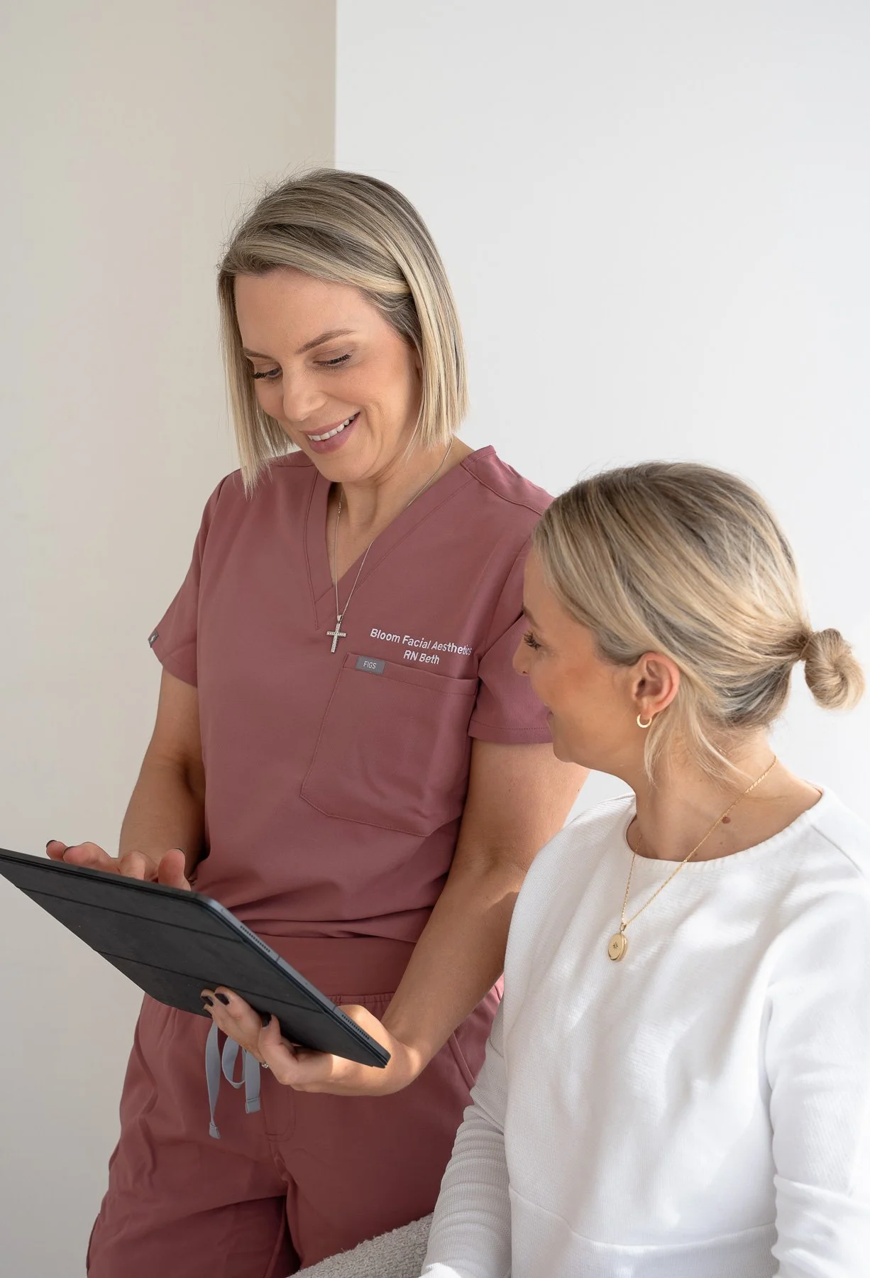 A healthcare professional in pink scrubs showing a tablet to a patient in white clothing, both smiling indoors.