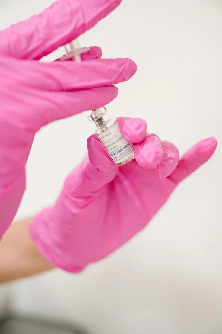 Person wearing pink gloves holding a small vial of vaccine or medication with a syringe ready for injection.