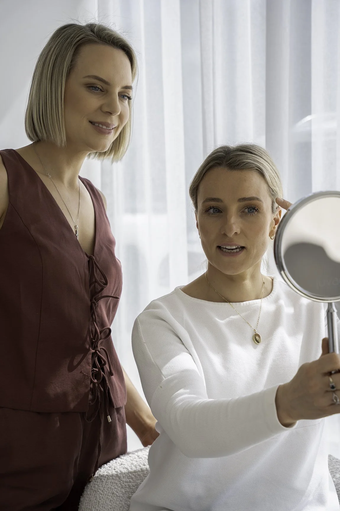 Two women, one seated and applying makeup, the other standing nearby in a well-lit room with white curtains.
