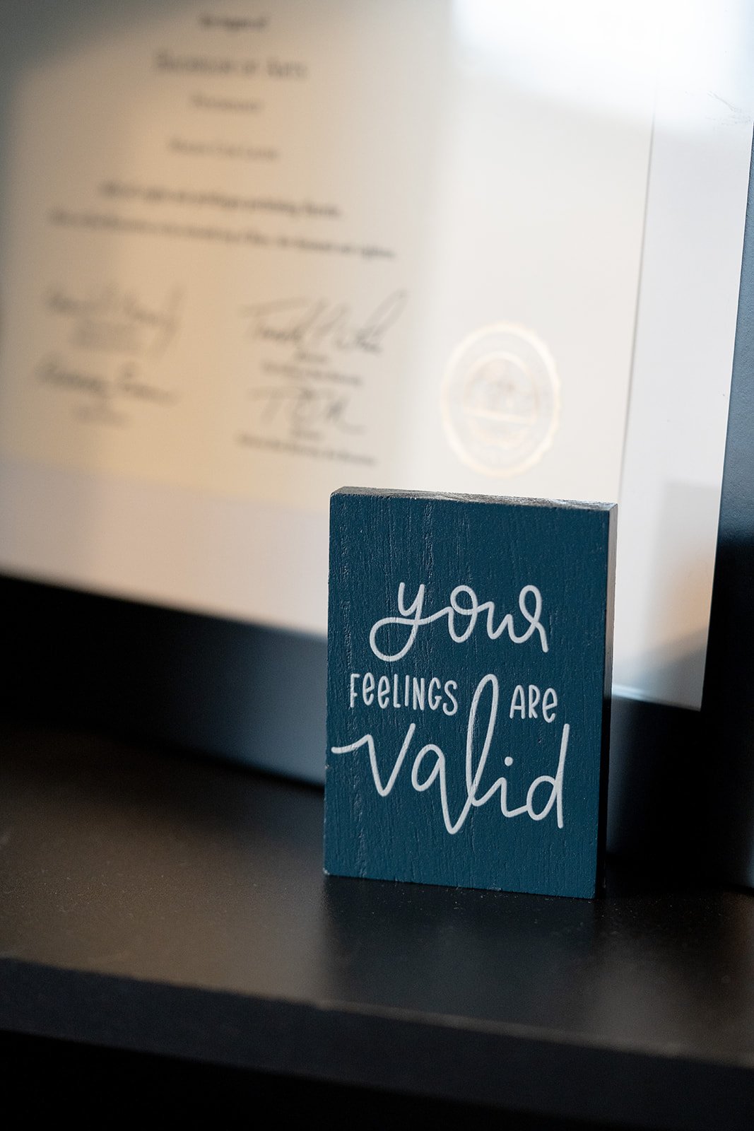 A blue wooden block with the words "your feelings are valid" in white lettering, placed on a black surface in front of a blurred certificate or diploma in the background.