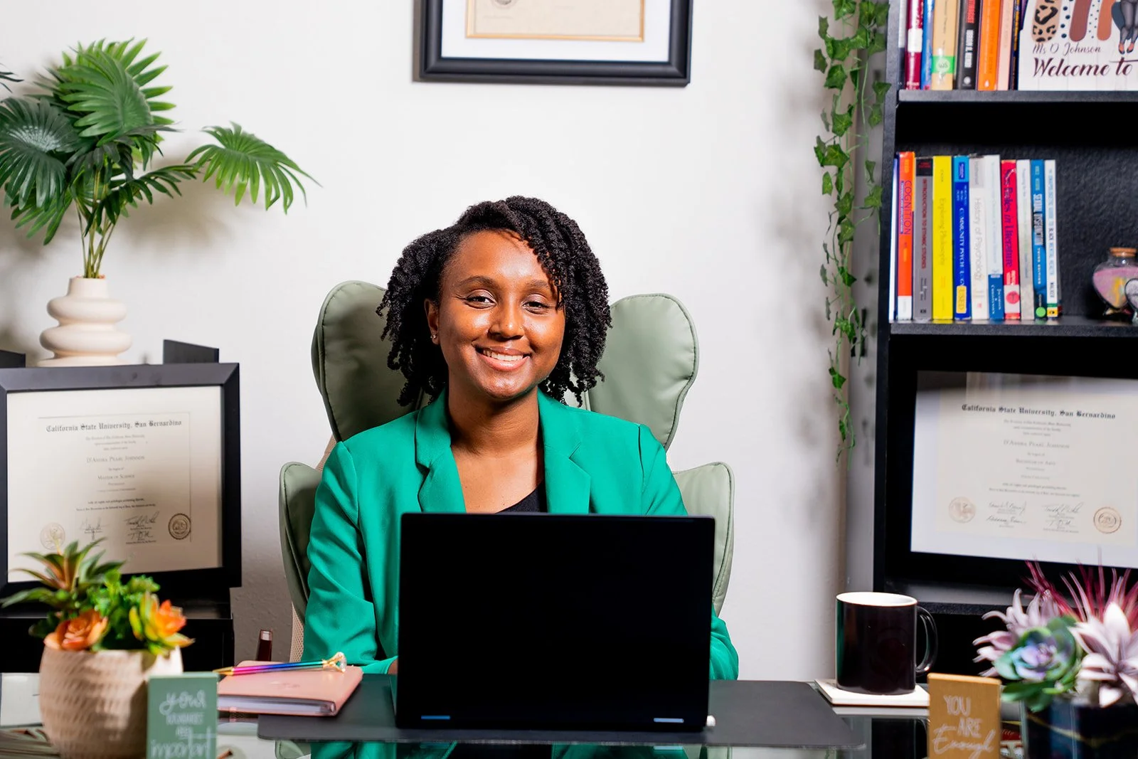 A brown skinned Black woman in a green blazer sitting at a desk with a laptop, surrounded by books, framed diplomas, and plants in an office setting.