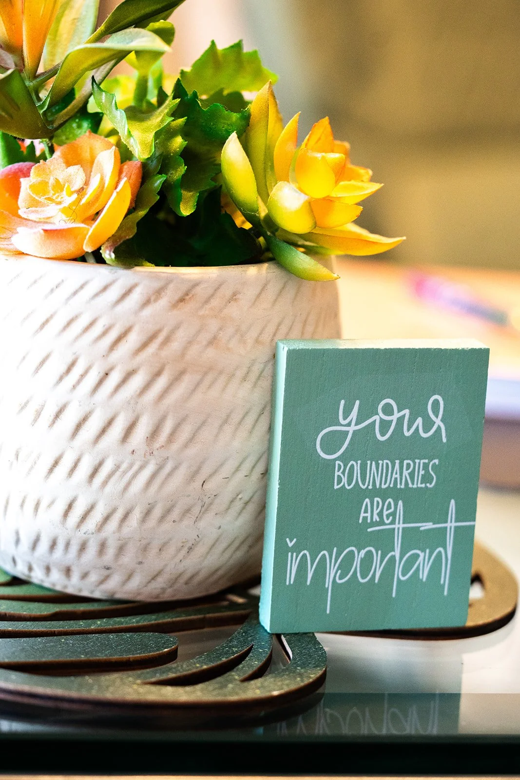 A white textured ceramic pot containing yellow and peach-colored artificial flowers and green foliage. Next to the pot is a small green sign with white handwritten text that reads, 'your boundaries are important.' The setup is on a decorative metal tray, placed on a reflective surface with a blurred indoor background.