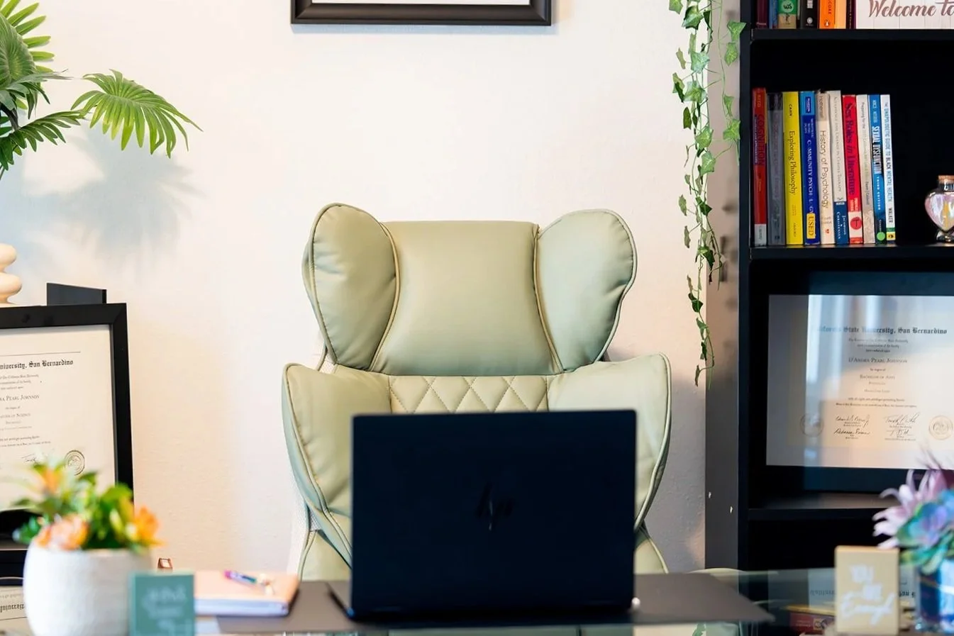 An office space with a beige high-back chair, a black bookshelf filled with books and certificates, and various decorative plants and items on the desk and shelves.