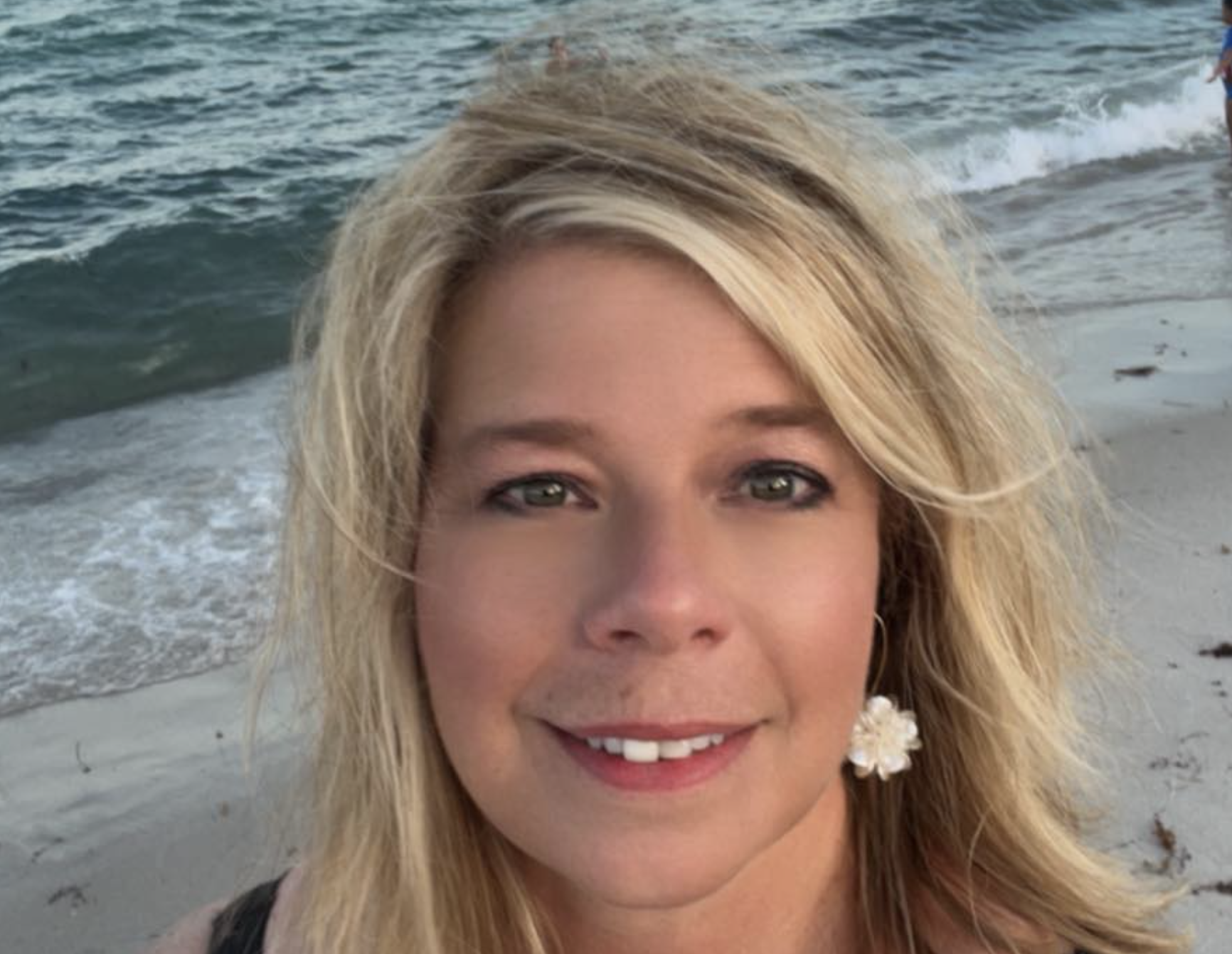 Close-up of a woman with blonde hair and green eyes smiling on a beach with ocean waves in the background.