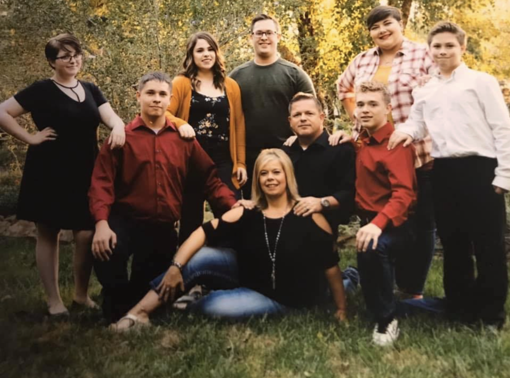 A group of nine people, including children and adults, posing outdoors in a park with trees and autumn leaves in the background.