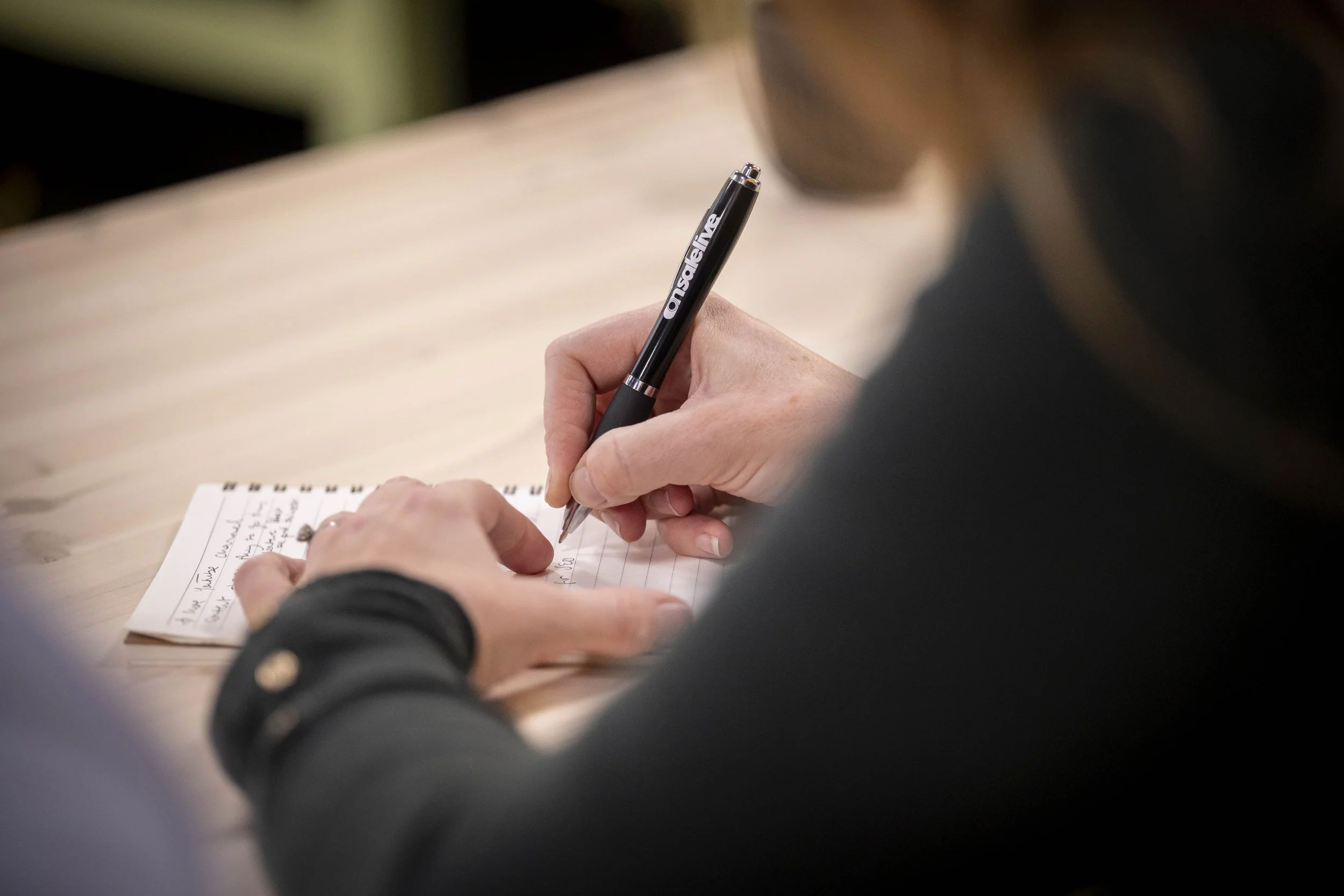 A person at GIEM arts, culture, events, experiences and live entertainment marketing course writing notes on a notepad with a black pen, wearing a dark long-sleeve shirt, on a wooden desk.