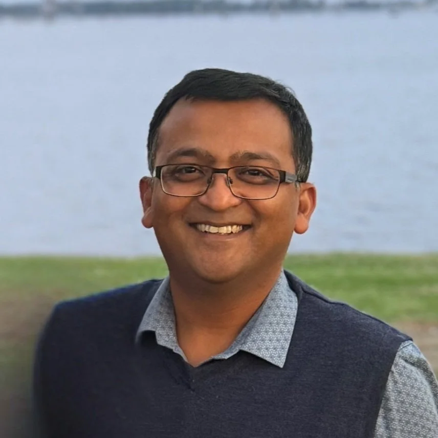A smiling man wearing glasses and a collared shirt standing outdoors near a body of water, with grass and water in the background.