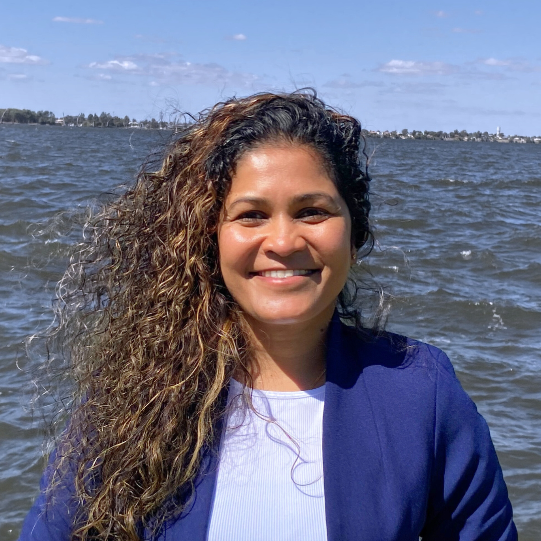 A woman with curly hair smiling outdoors near water, with a cityscape in the distance.