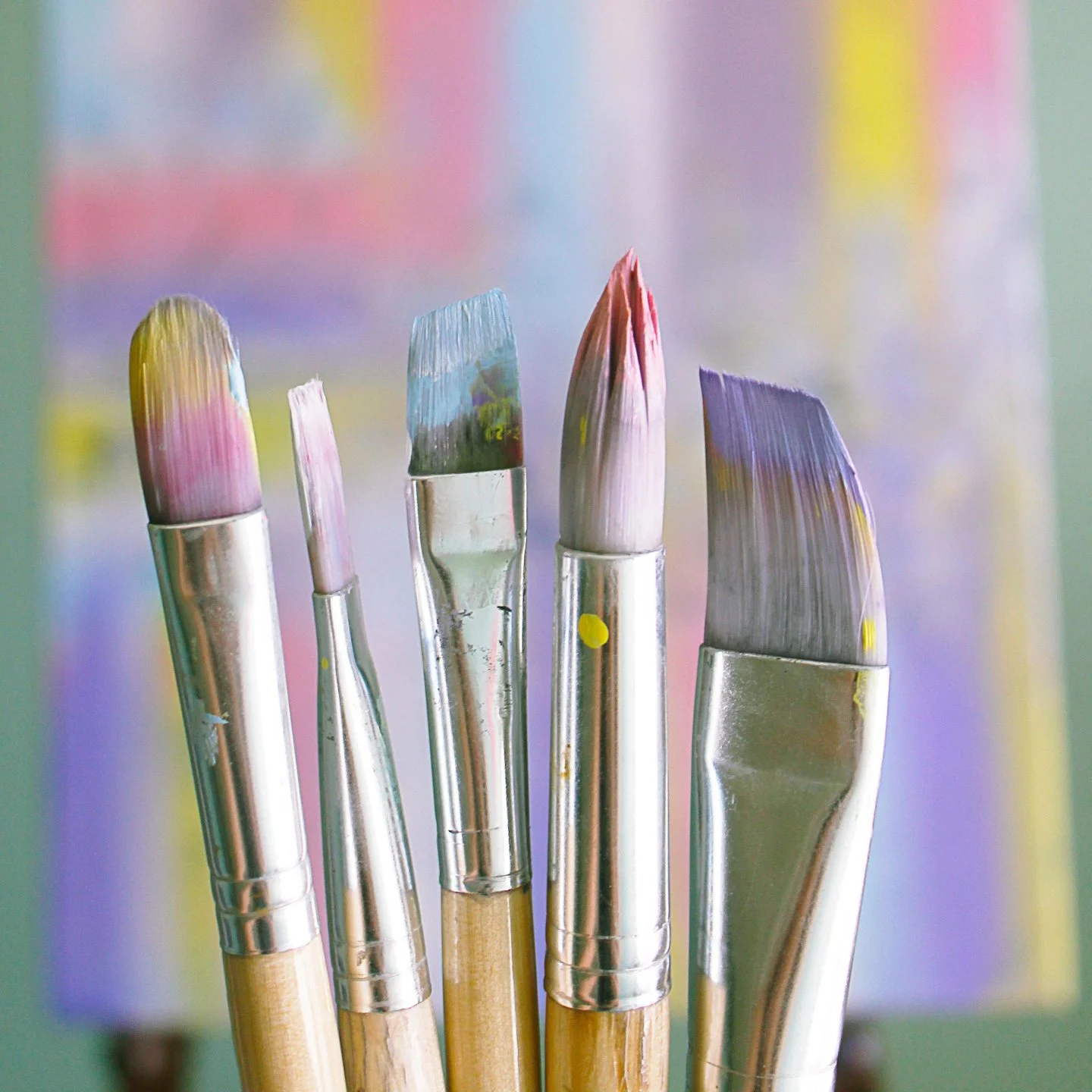 Close-up of five paintbrushes with colorful paint on their bristles, standing upright against a blurred colorful background.