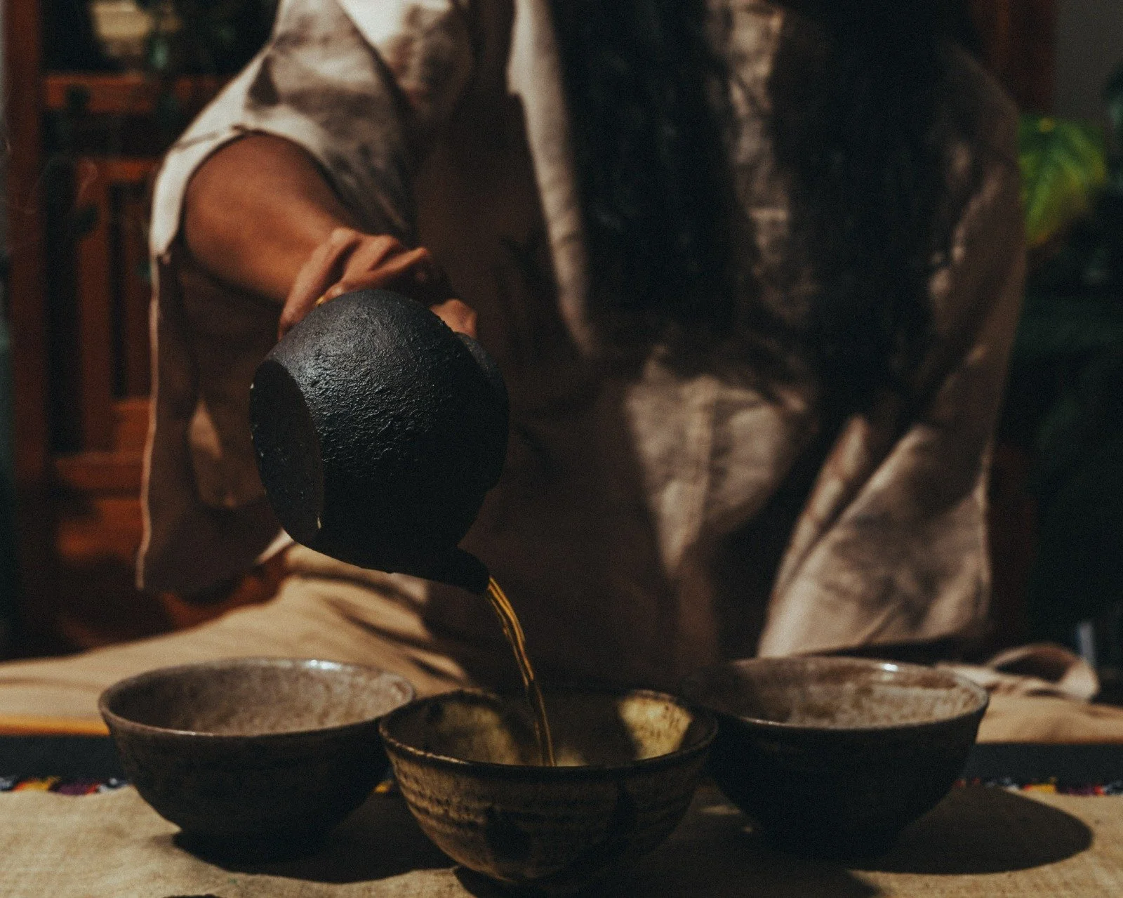 A woman with closed eyes, wearing a light-colored top and a delicate necklace, standing outdoors with sunlight on her face and dark blurred lush greenery in the background.