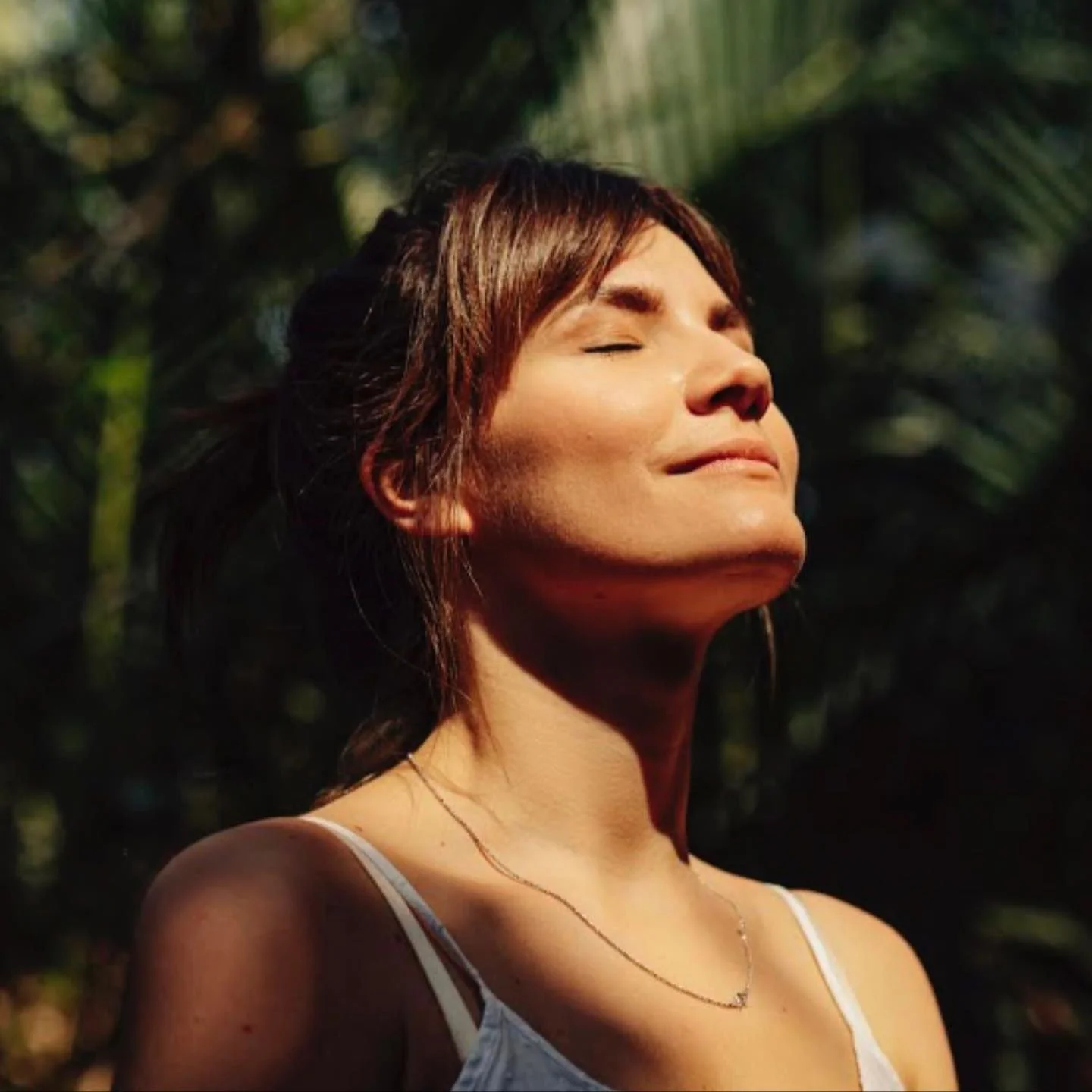 A woman with closed eyes, wearing a light-colored top and a delicate necklace, standing outdoors with sunlight on her face and dark blurred lush greenery in the background.