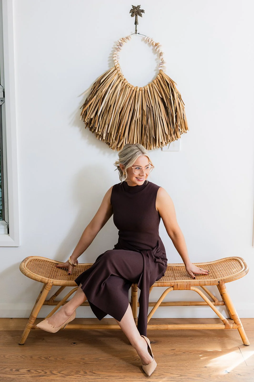 A woman with glasses and blonde hair styled in a bun, wearing a sleeveless dark brown dress and beige high heels, sitting on a wooden bench with her legs crossed, smiling and looking to the side. Behind her, a large, decorative wall hanging made of natural materials, resembling a circular frame with a fringe of tan-colored palm leaves, is mounted on a white wall.