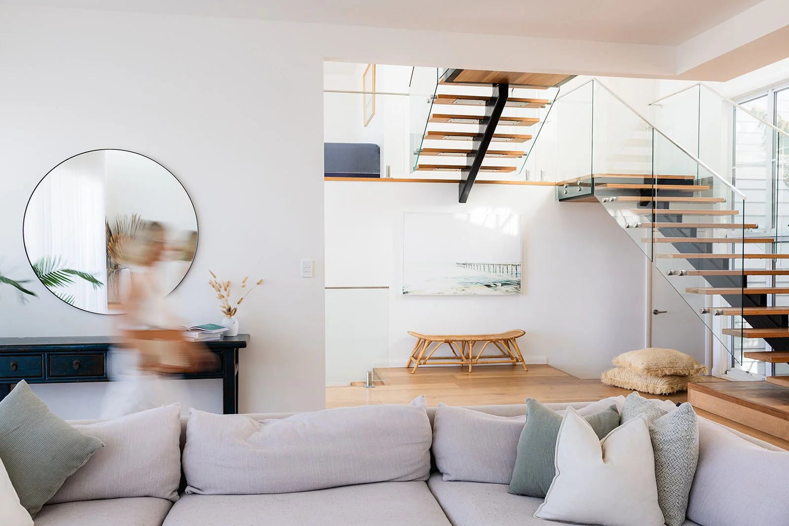 Modern living room with a white couch, decorative pillows, a round mirror, a console table with a plant and books, a staircase with wooden steps and glass railings, a wall art piece, and minimalist decor.