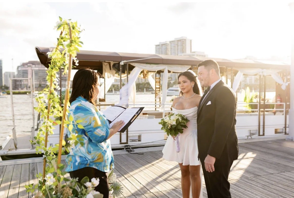 A group of people dining on a boat at sunset, with servers serving food and drinks, and a city skyline in the background.