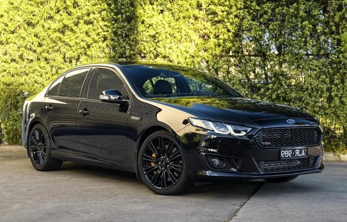Black sedan car parked on a concrete surface with green bushes in the background.