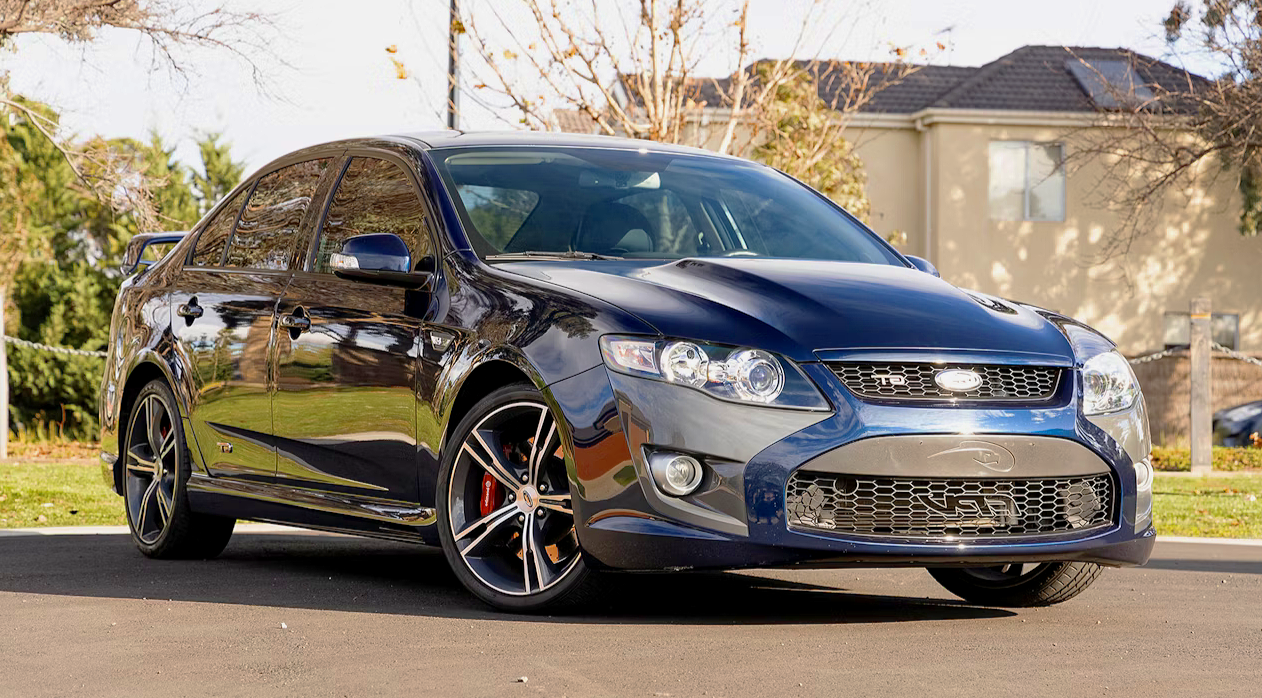 Black sedan parked on a driveway with residential houses and trees in the background.