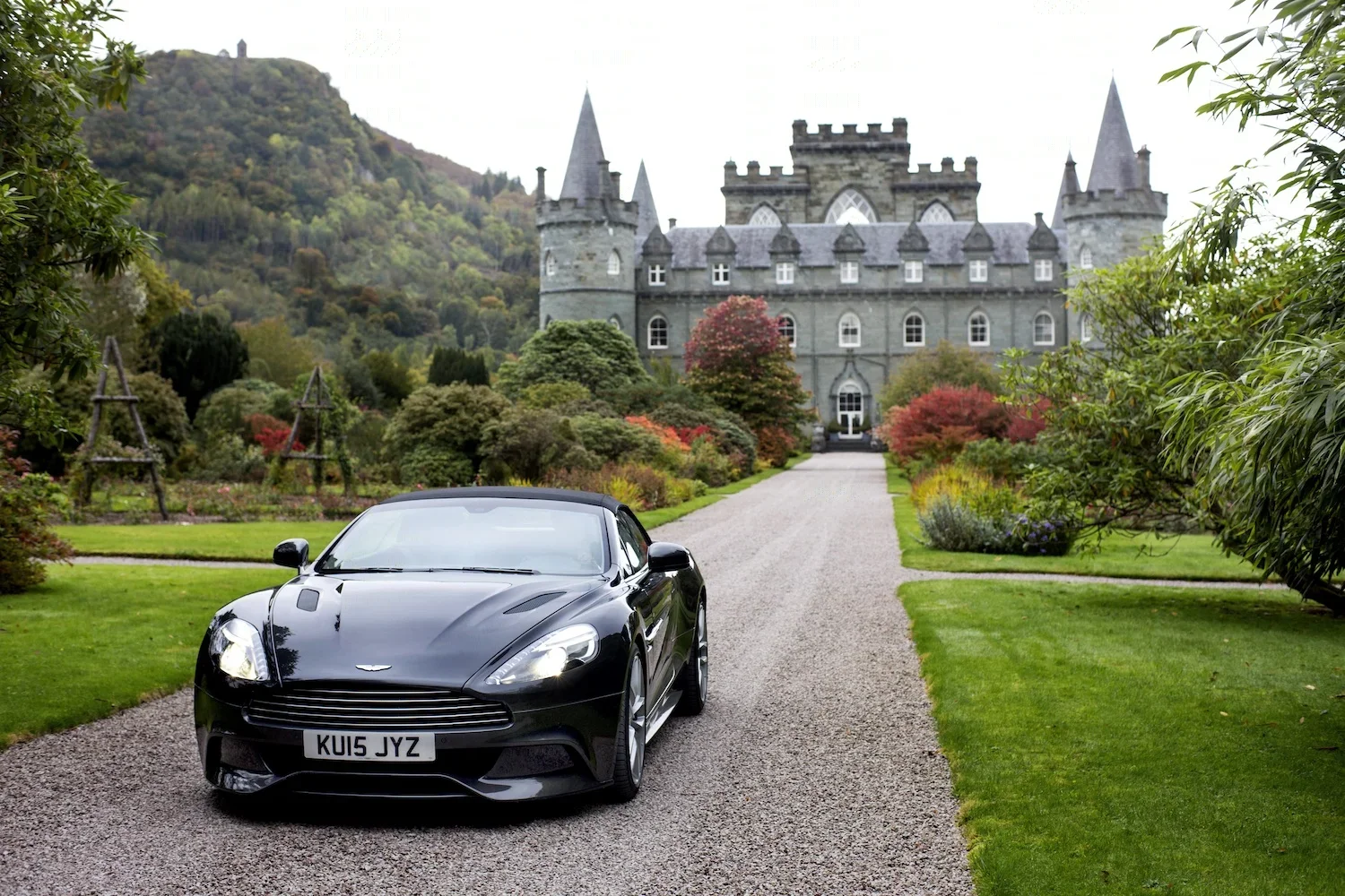 A black Aston Martin sports car parked on a gravel driveway in front of a large gray castle with multiple towers, surrounded by colorful autumn trees and well-maintained gardens.