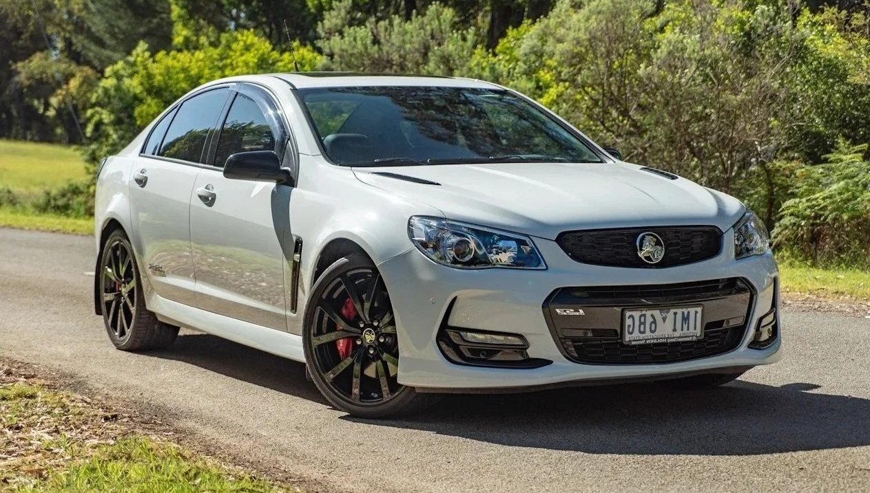 A white sedan car parked on the side of a rural road with trees and greenery in the background.