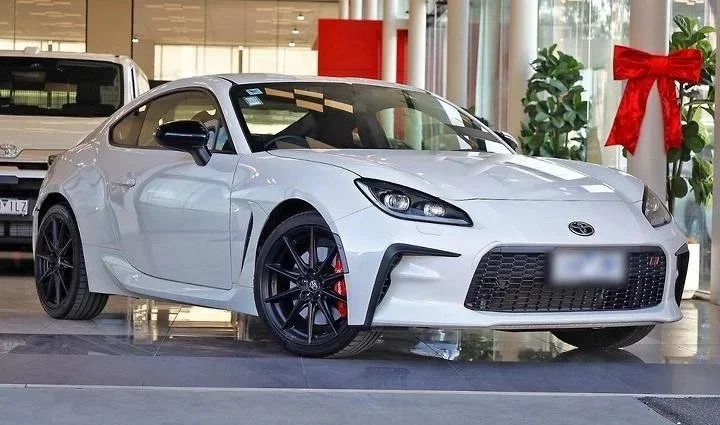 A white Toyota sports car with black wheels and red brake calipers, displayed inside a dealership with a large red bow decoration nearby.