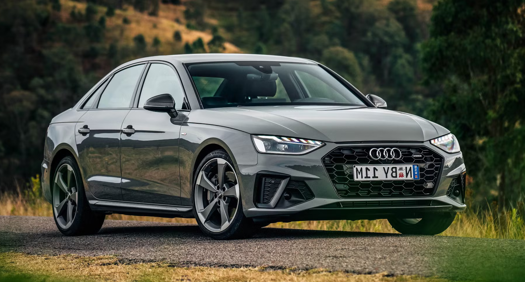 A silver Audi sedan parked outdoors on a road with green trees and hills in the background.