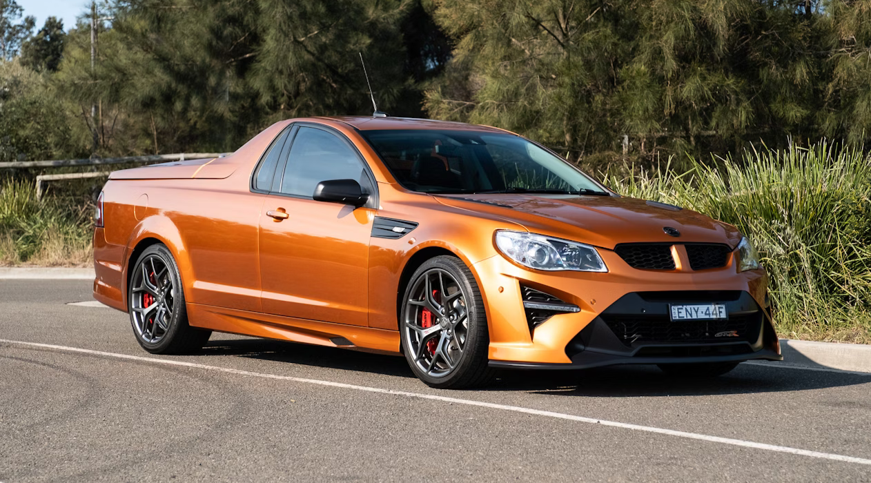 An orange sporty coupe parked on the side of a road with trees and greenery in the background.