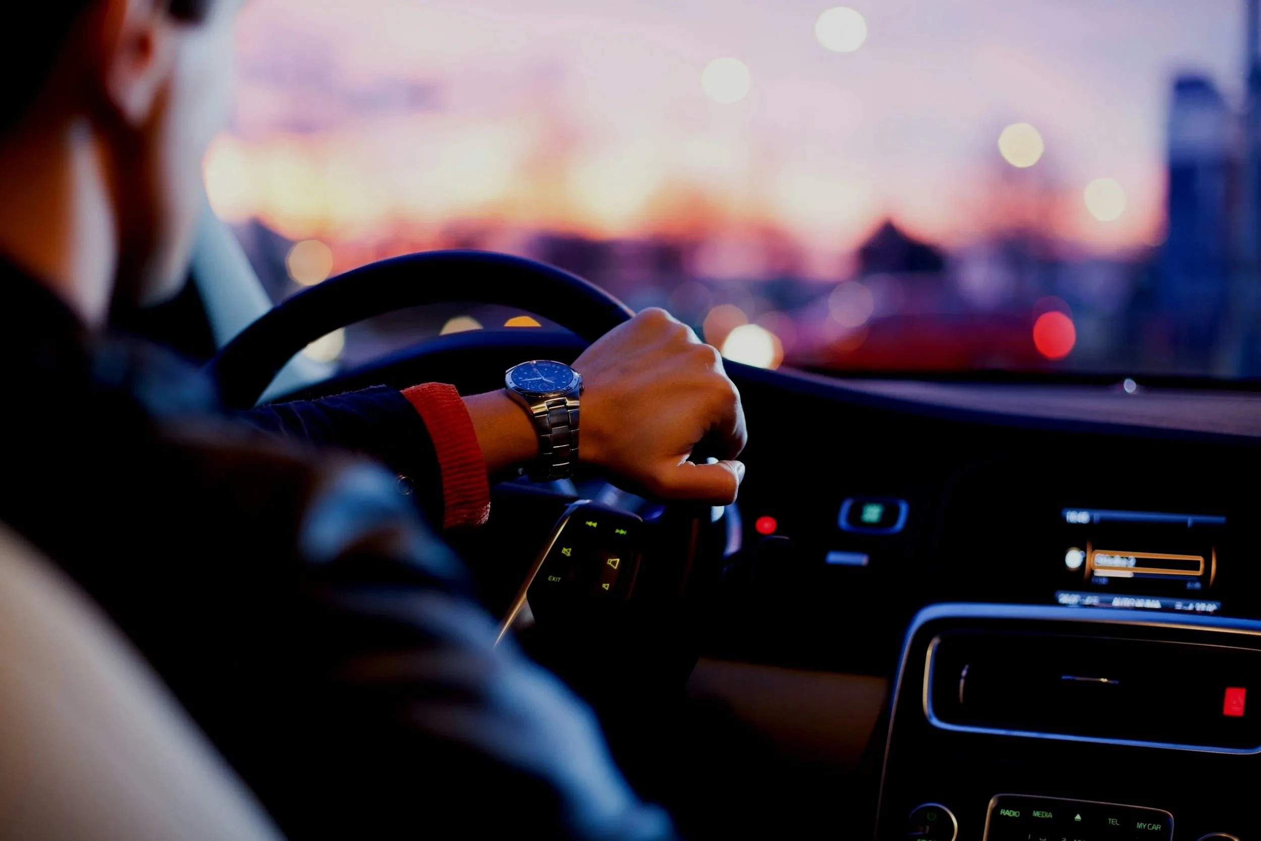 Person driving a car during sunset, wearing a watch on their left wrist, with a blurred cityscape in the background.