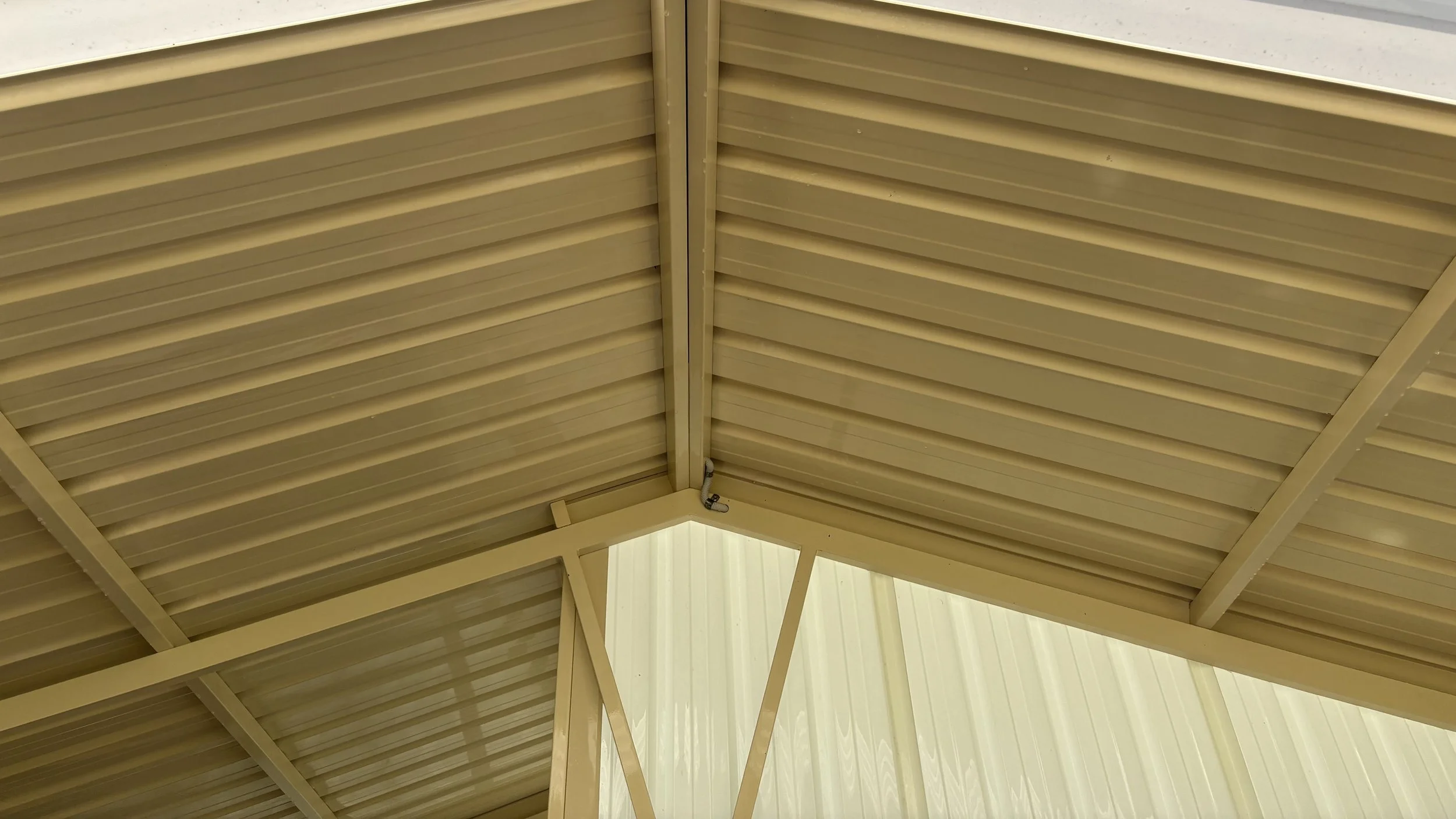 Close-up view of a beige metal roof with overlapping panels, featuring beams and a corner with a small pipe or conduit attached.