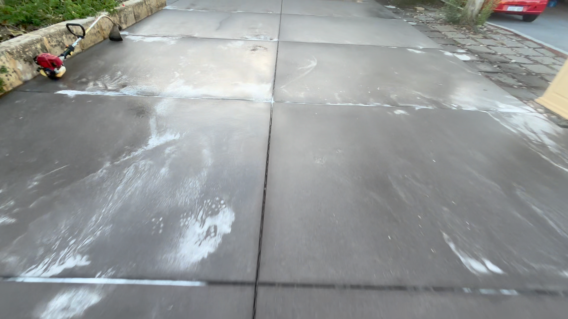 Freshly sealed or cleaned concrete sidewalk with a few cleaning tools and soap suds visible, bordered by a flower bed and a street with parked cars.