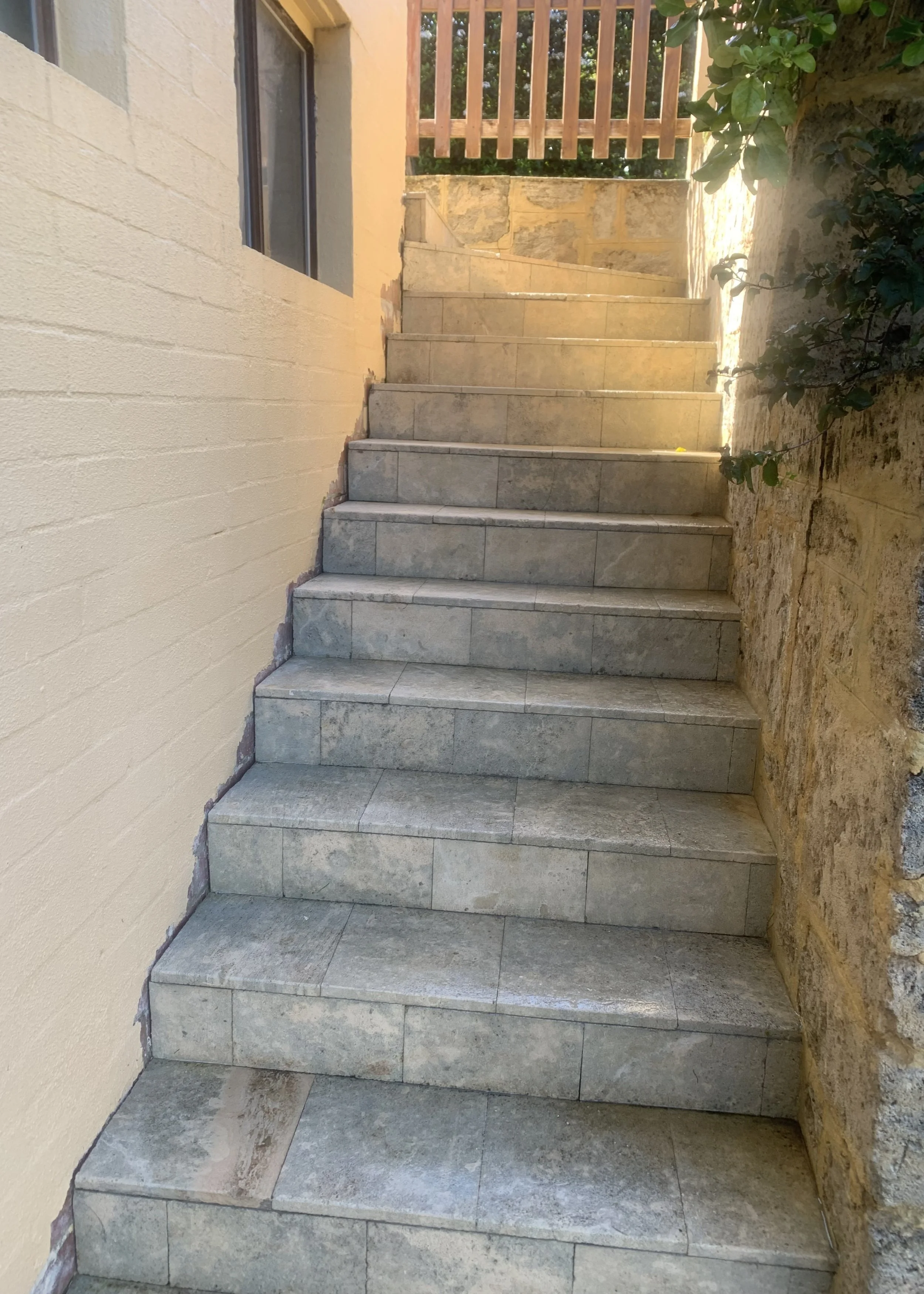 Concrete stairs with a wooden fence at the top, flanked by a brick wall on the left and a stone wall on the right, with some green plants on the right side.