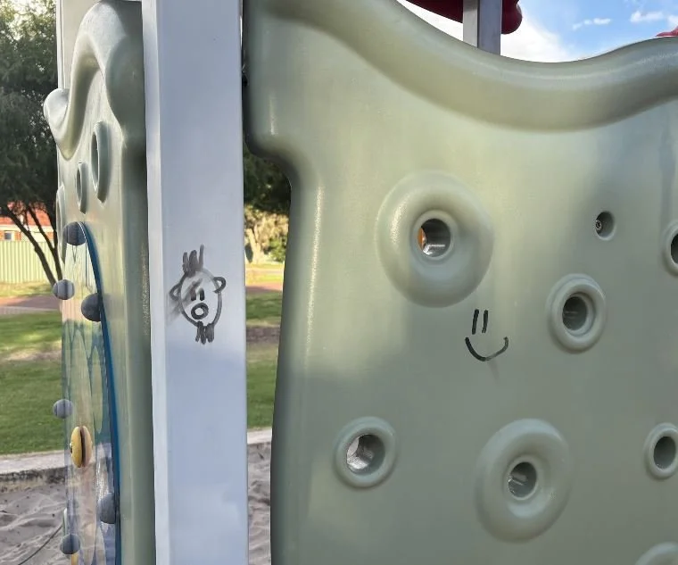 Close-up of a playground climbing wall with handholds, featuring drawings and a smiley face on its surface, outdoors with grass and trees in the background.