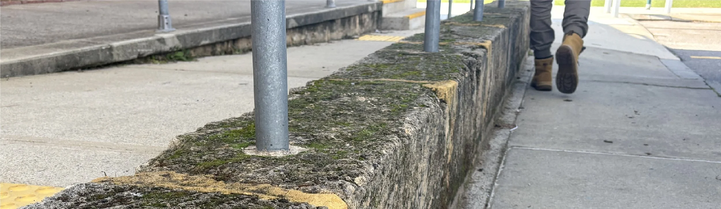 Sidewalk with moss-covered concrete barrier, metal poles, and a person walking away wearing brown boots and dark pants.