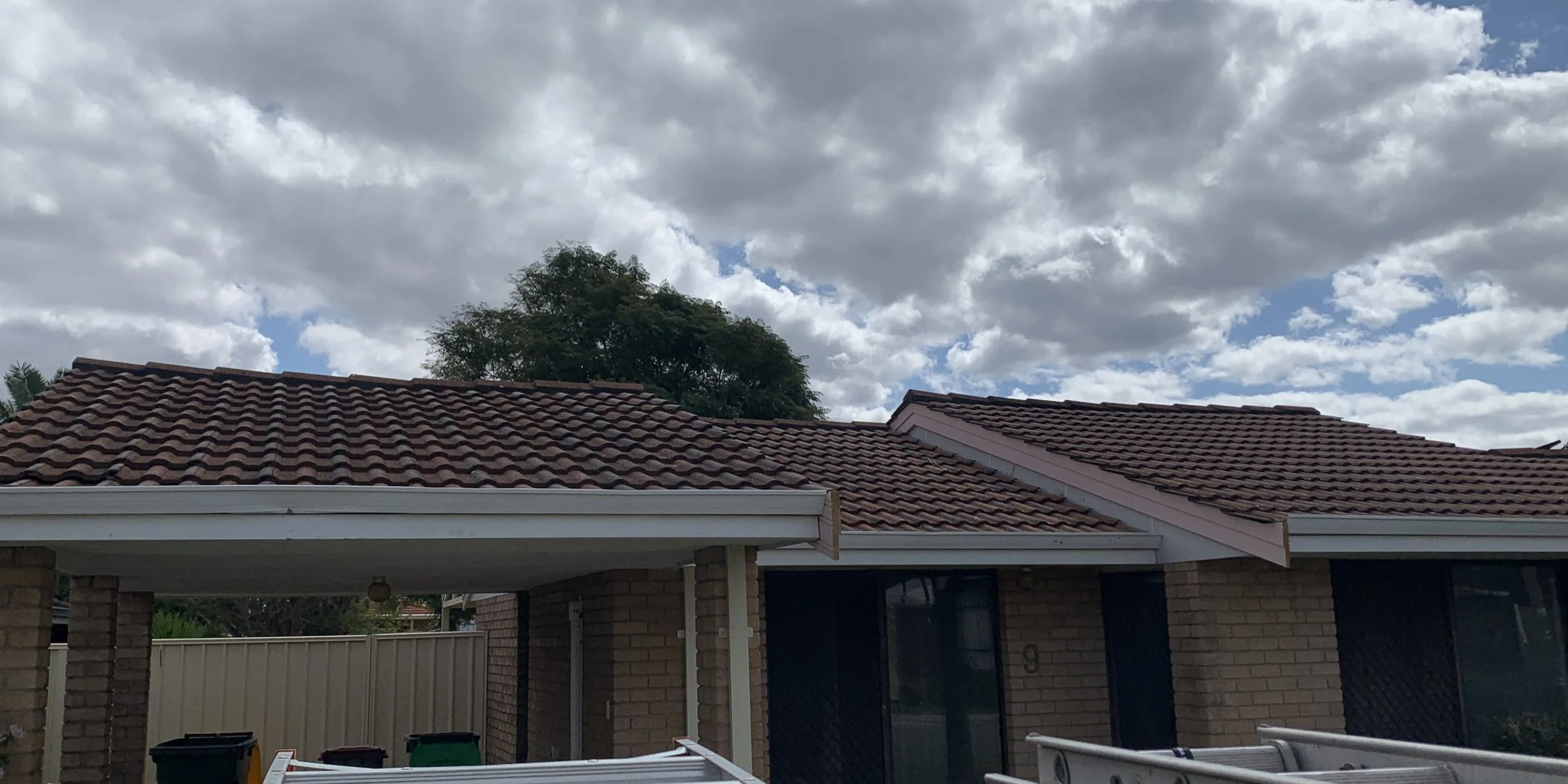 View of a house with a tile roof, brick walls, a covered porch, and a backyard with a fence and garbage bins. The sky is partly cloudy with patchy blue sky visible.
