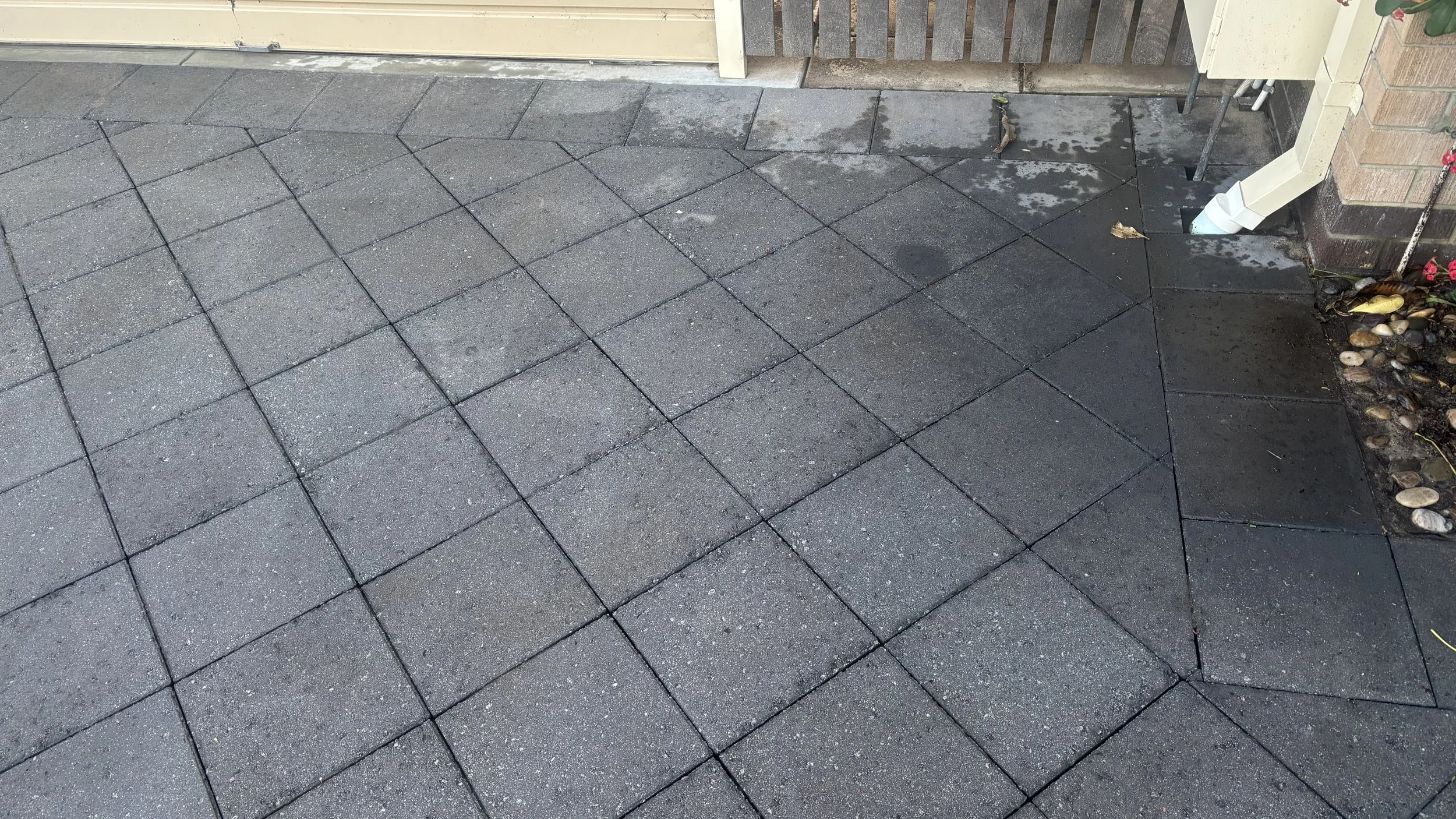 Concrete patio with a grid of square pavers, some wet spots, and a small flower bed with rocks and plants on the right side.