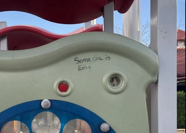 A close-up of a pale green plastic playground panel with a blue circular section and metal bolts. There is handwritten text on the panel that says, "Someone is Emo." In the background, part of a red slide and white metal structure are visible.
