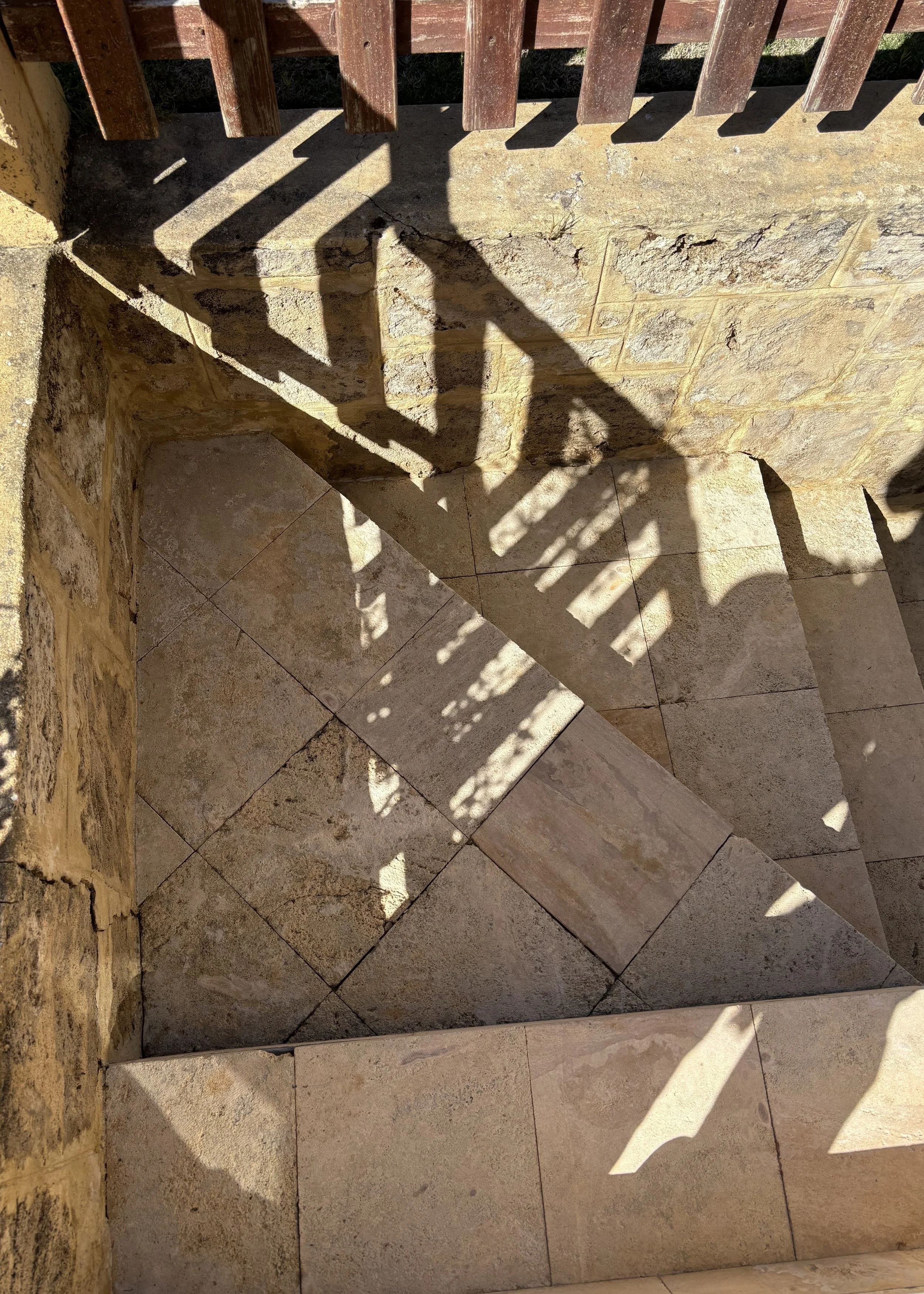 Shadows of a wooden fence on stone tiles and brick wall, with sunlight creating patterns on the ground.