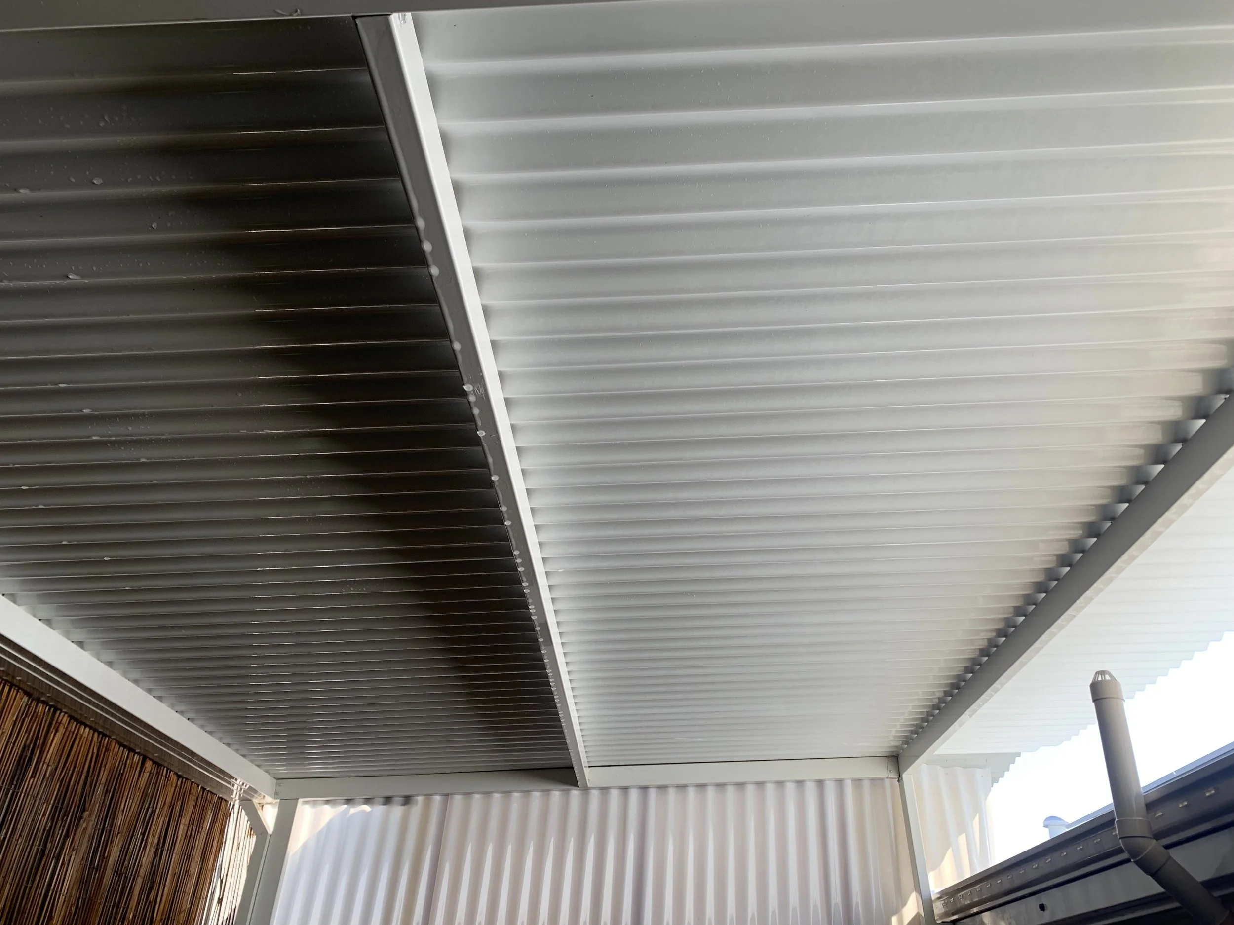 View of corrugated metal roof panels from below, showing part of a gutter and bamboo fencing on the side.