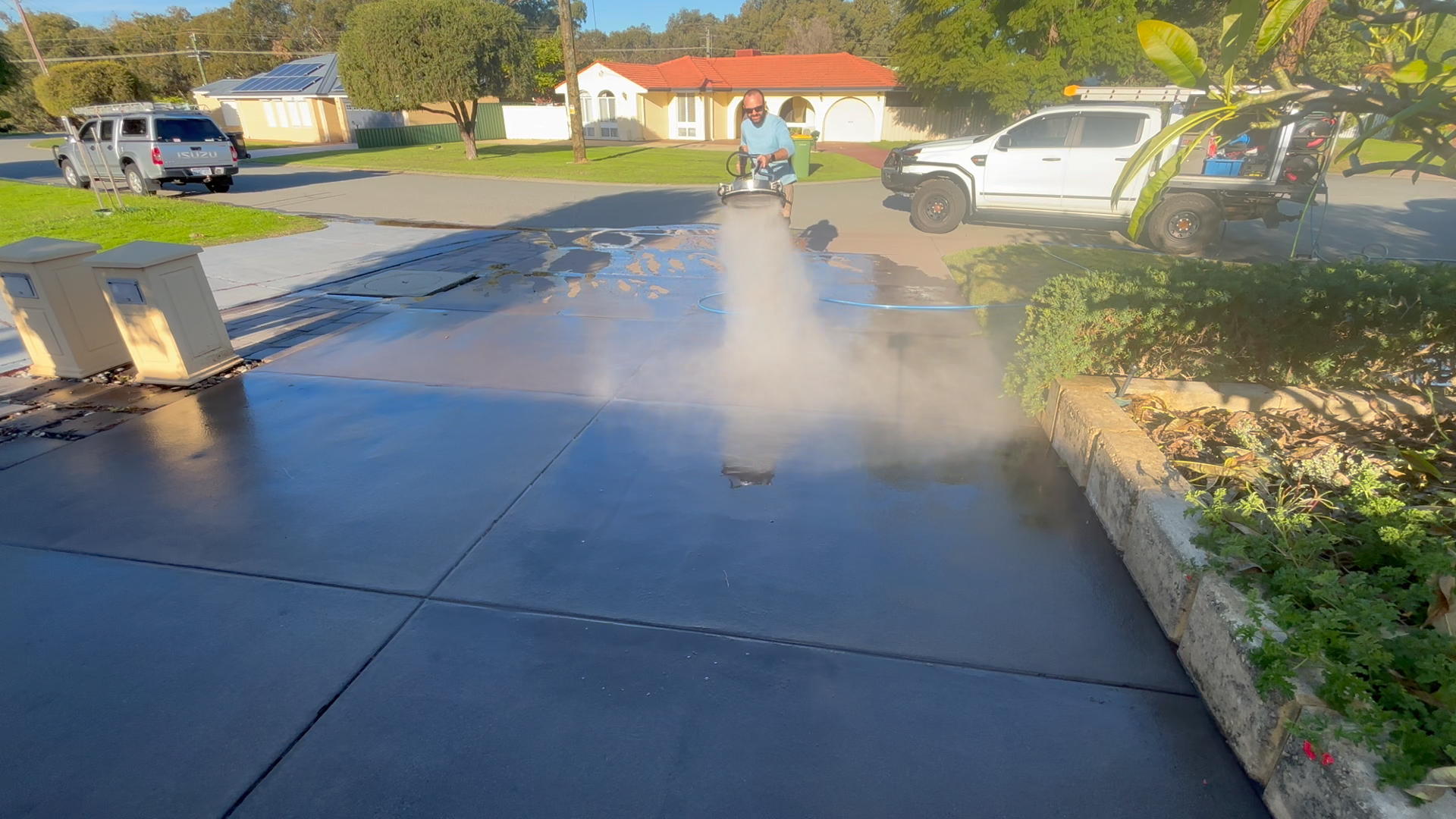 A man in sunglasses using a power washer to clean a concrete driveway outdoors in a suburban neighborhood with houses, trees, and parked trucks visible in the background.