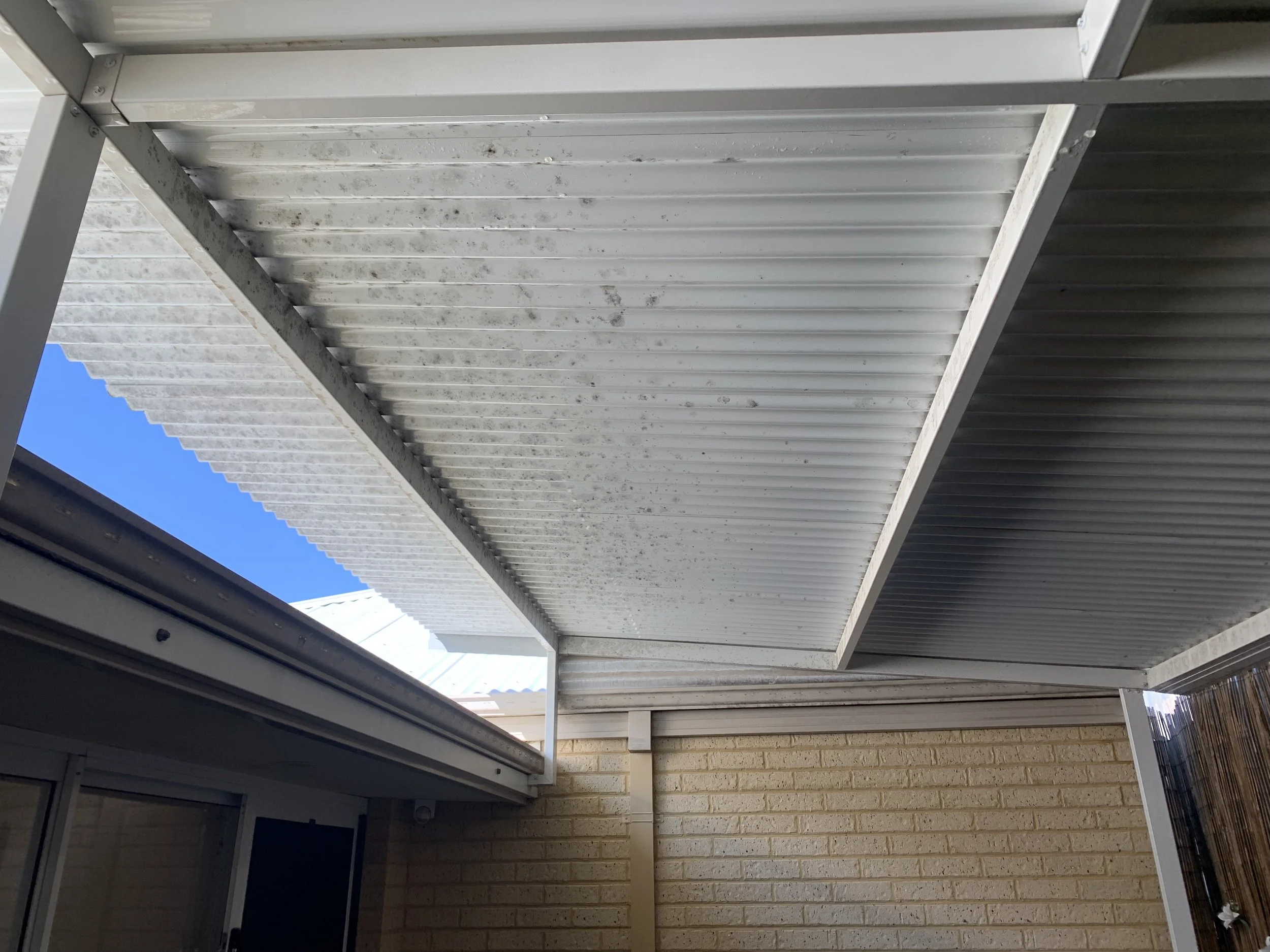 View of a corrugated metal roof with a small opening revealing blue sky, viewed from below, attached to a brick building with a window.