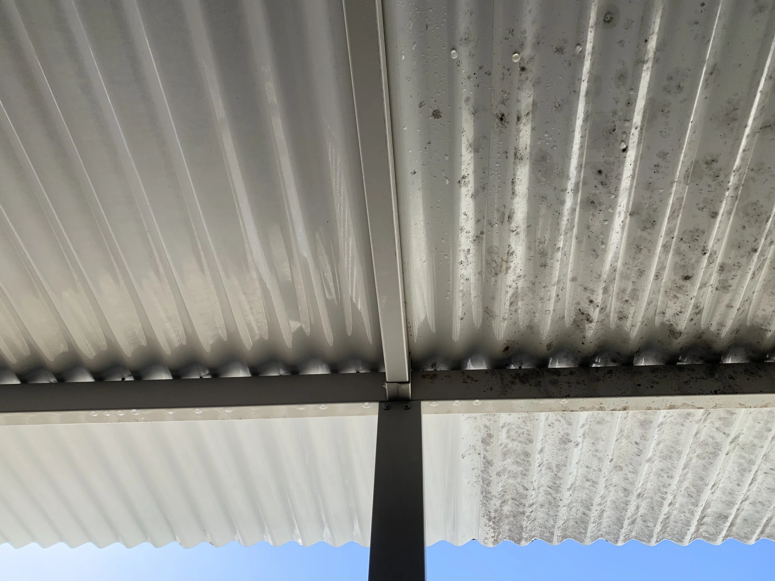 Looking up at a divided metal roof with visible dirt and stains, supported by a black pole, against a clear blue sky.